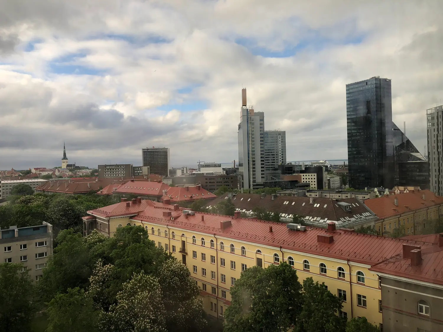 Roofs, towers and a spire of an old town seen from above, with trees and a tall block beyond them.