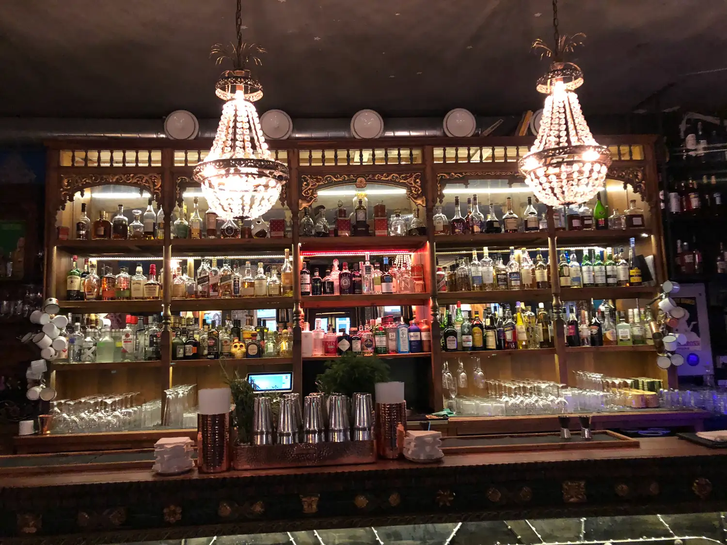 A bar back of shelves and bottles with two chandeliers above it.