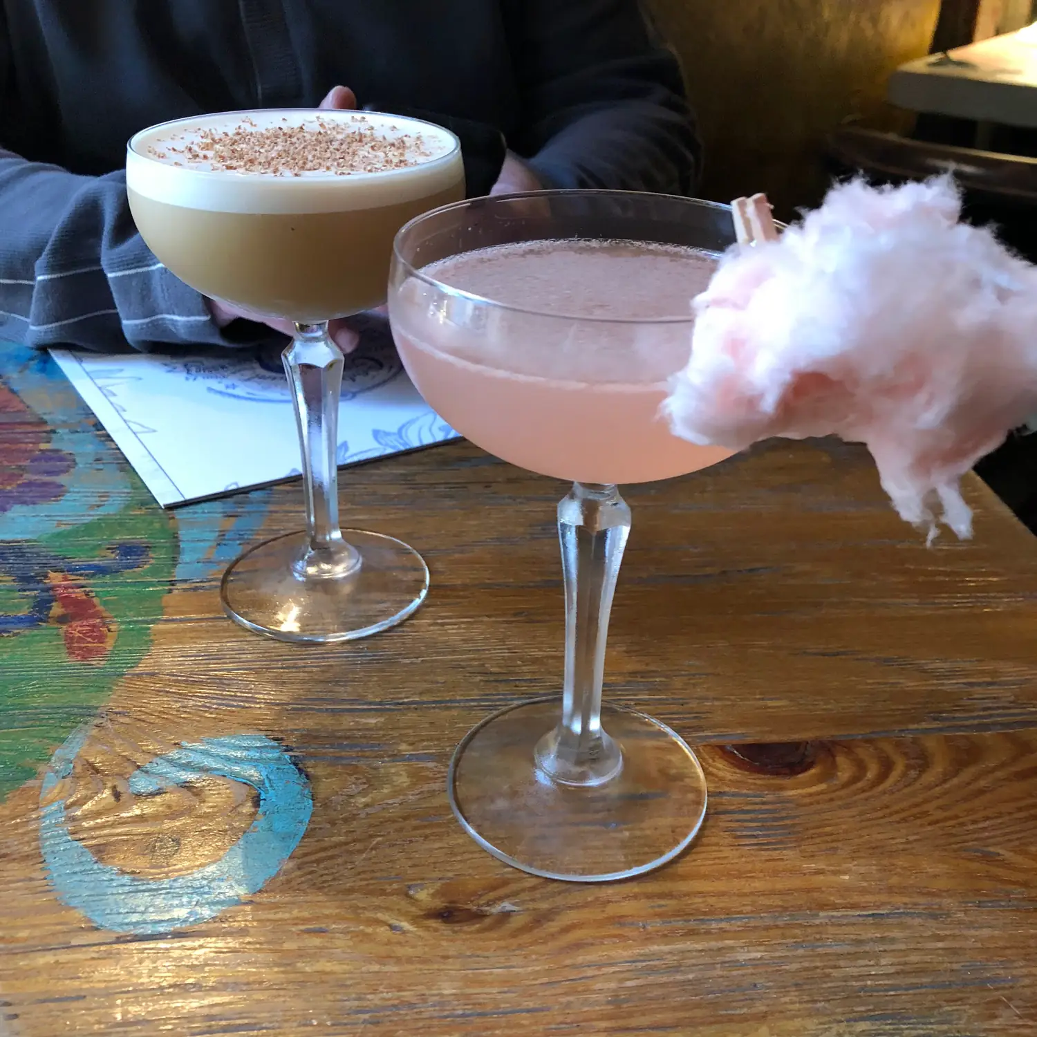 Two cocktails seen from above on a table, one tall and pale with foam on top and one pink in a stemmed glass, with a person's hands and sleeve at the top edge.