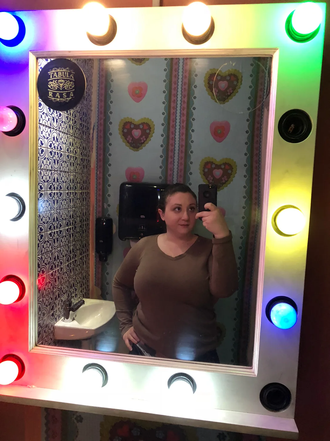 A woman taking a picture of herself in a mirror framed with round lights, in a room hung with pictures.