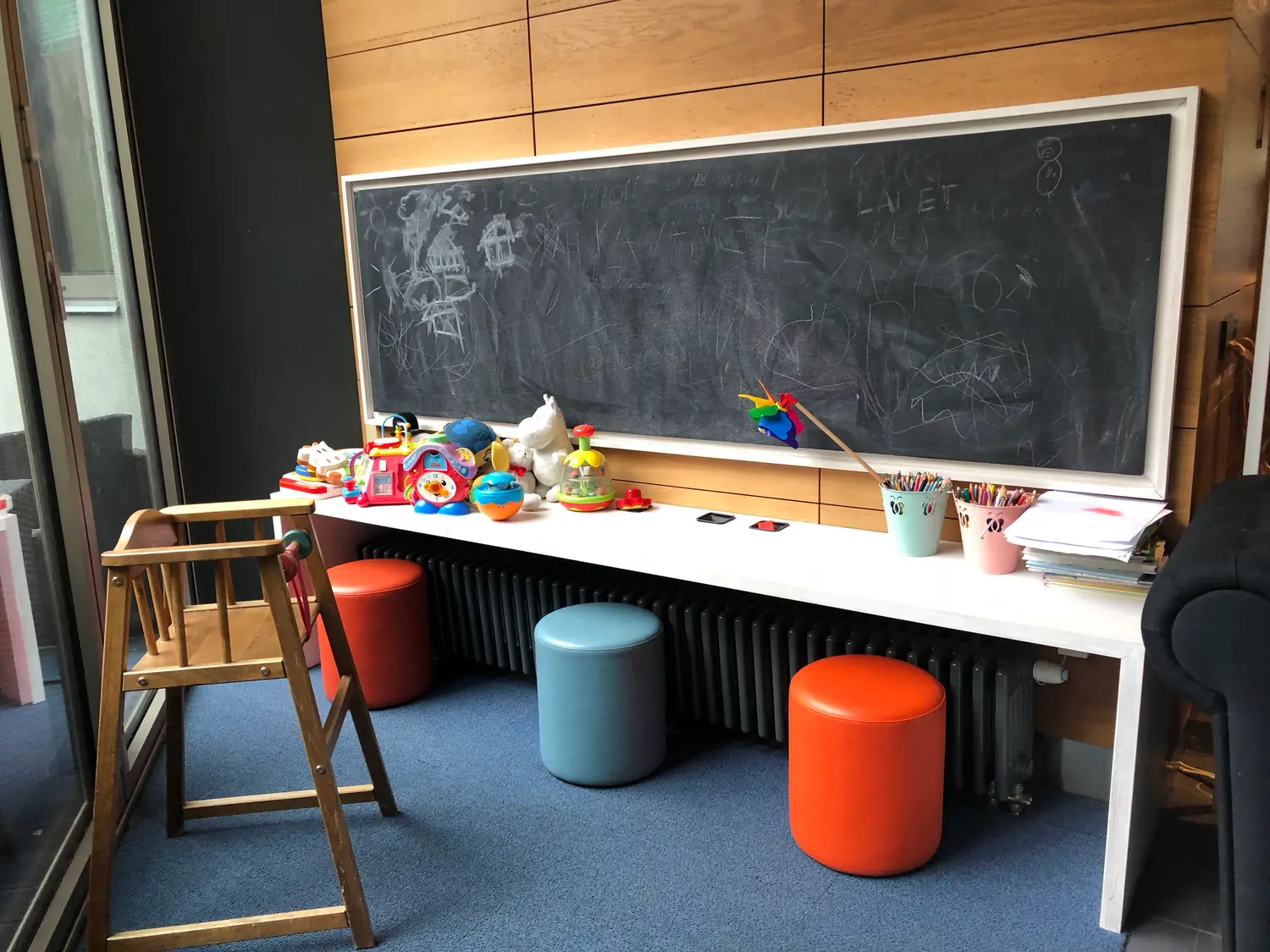 A play corner with a blackboard along the wall, a low bench carrying small toys, and a chair and two stools on a blue floor.