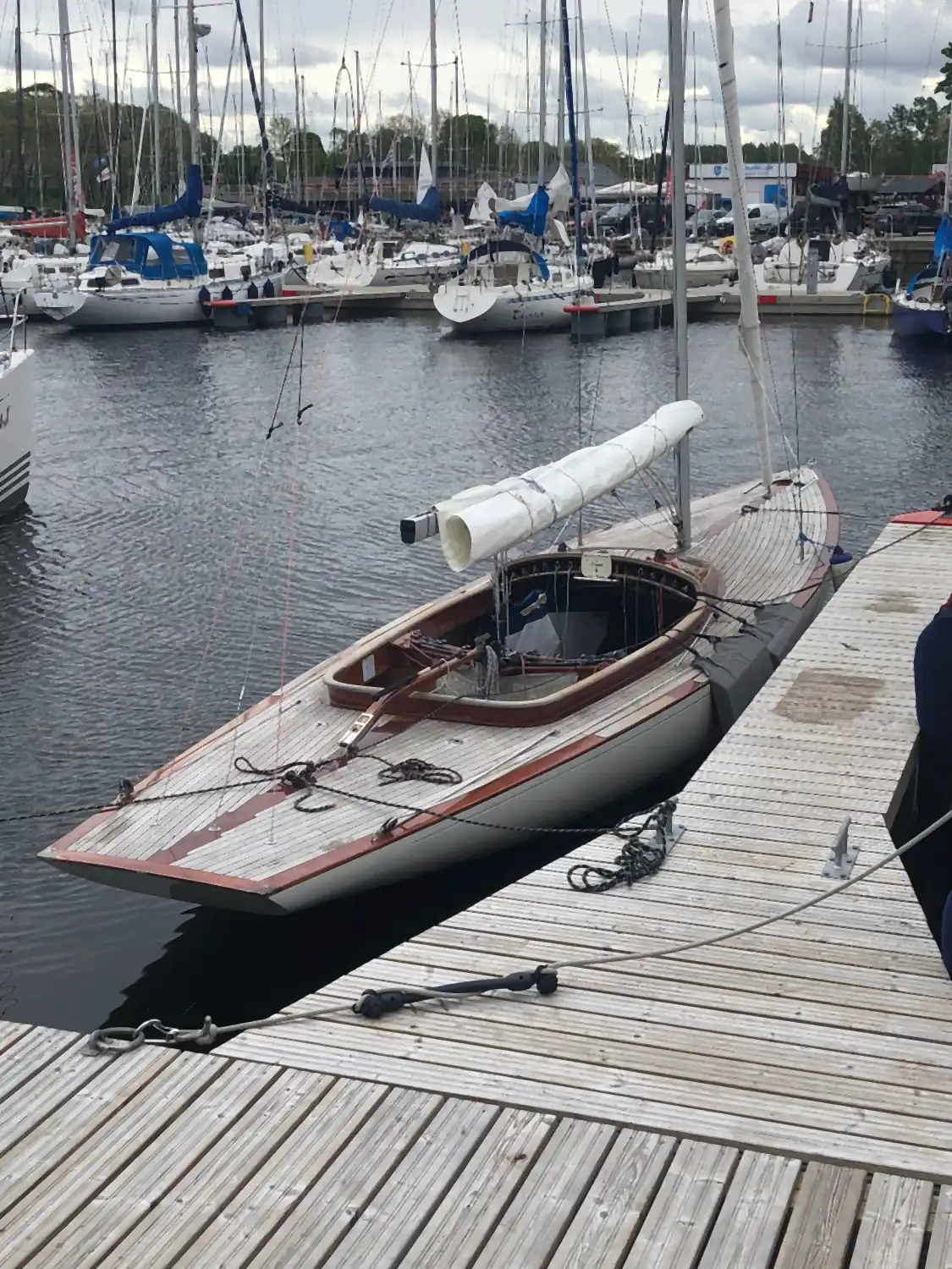 A varnished wooden launch moored along a timber pontoon, with masts and other boats behind it.