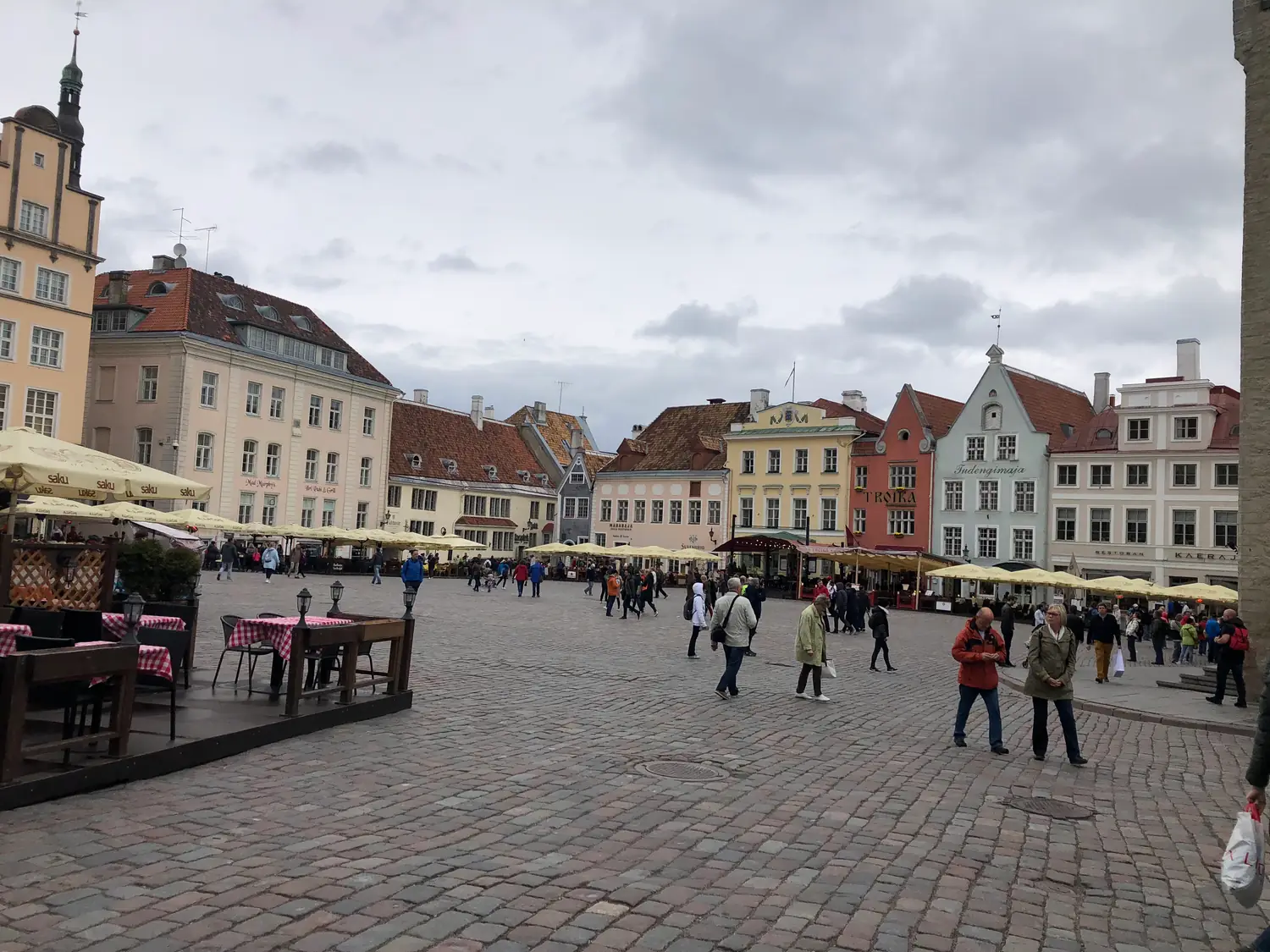 A town square with market umbrellas and a few people at a distance, old houses along the far side.