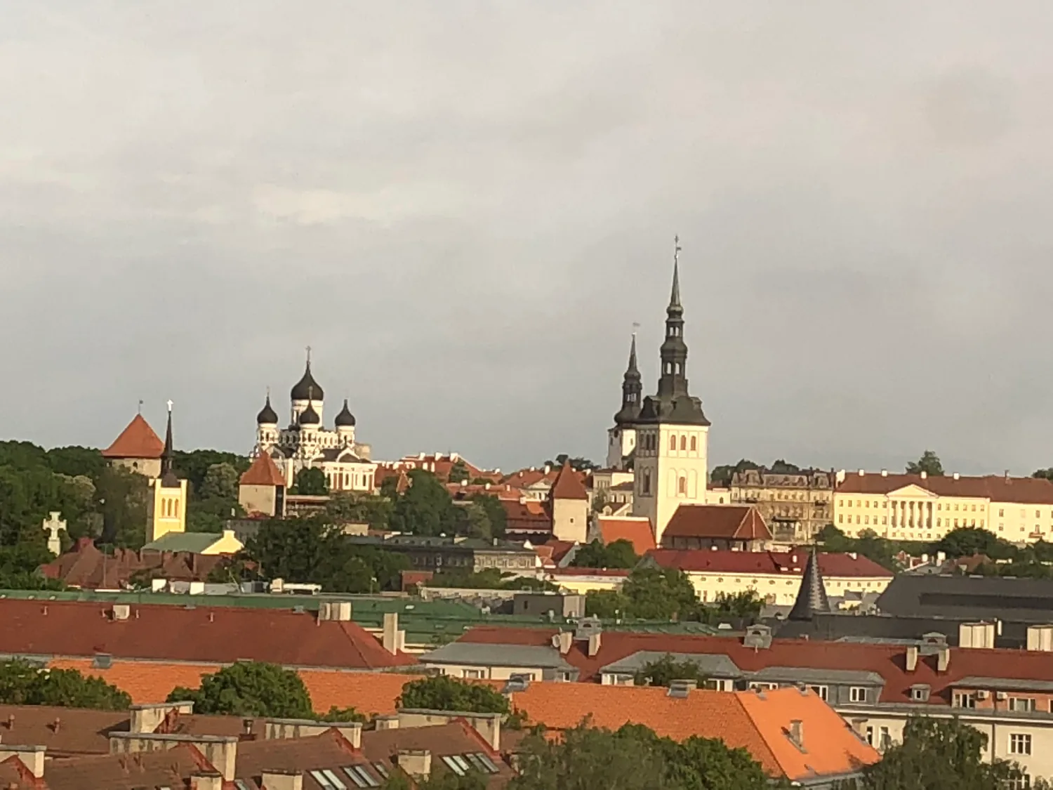 An old town skyline seen across the rooftops below: a slender spire and a tall tower rising over red tiled roofs, with dark domed cupolas further off and a wide pale sky behind them.