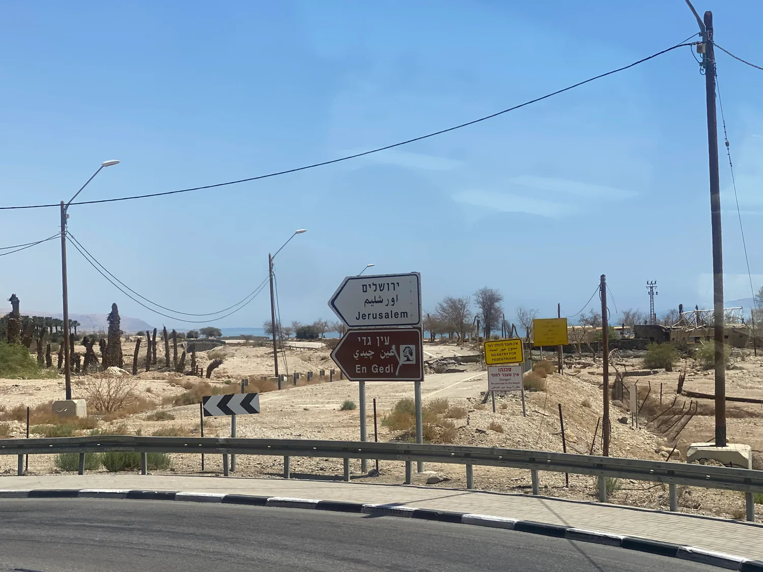 A junction in bare desert with road signs on a pole, a guard rail along the road in front and dry hills far behind.