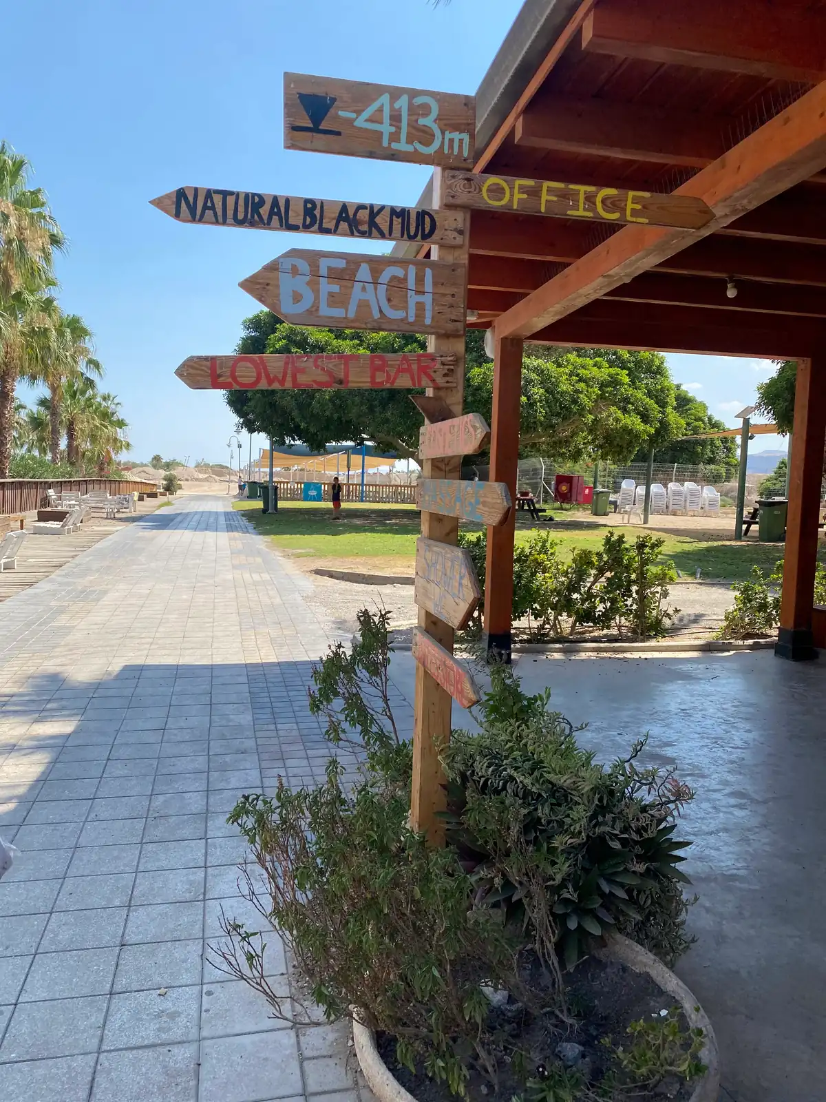 A wooden signpost of painted arrows pointing to the mud, the beach, the office and the lowest bar, under the roof of a pergola with a paved path behind it.