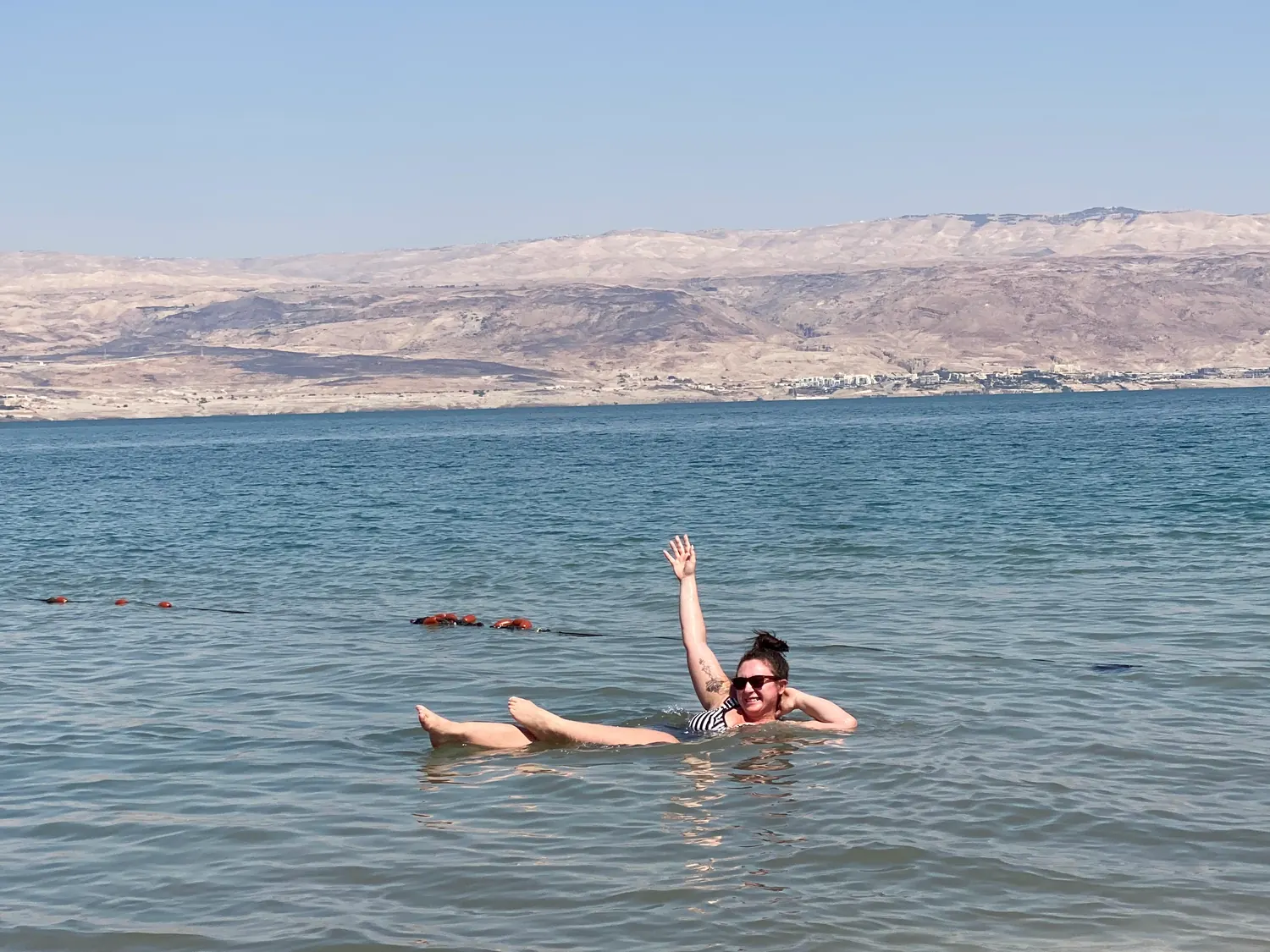 A woman in a striped swimsuit floating in dark blue water with one arm raised, bare hills along the far shore behind her.