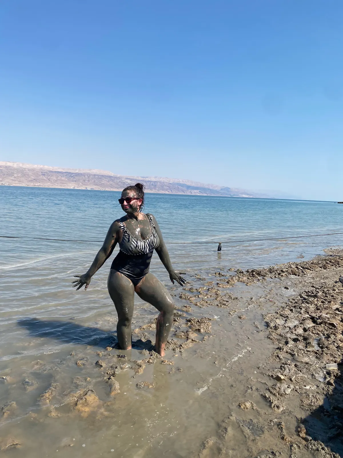 A woman standing at the edge of the water covered in grey mud from her shoulders down, with bare hills across the water behind her.