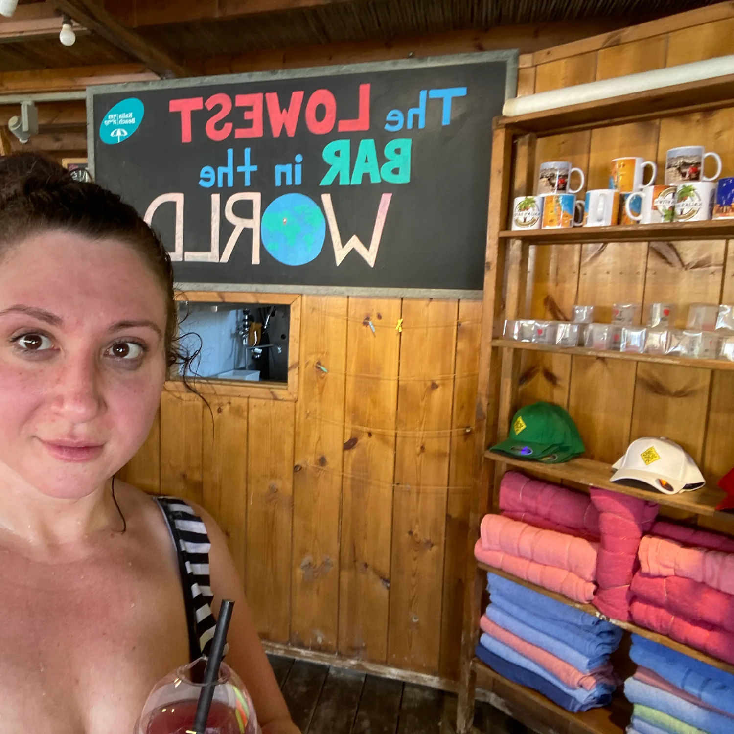 A selfie of a woman in a beach shop with a painted sign on the wall behind her reading the lowest bar in the world, folded towels stacked on the shelves beside her.
