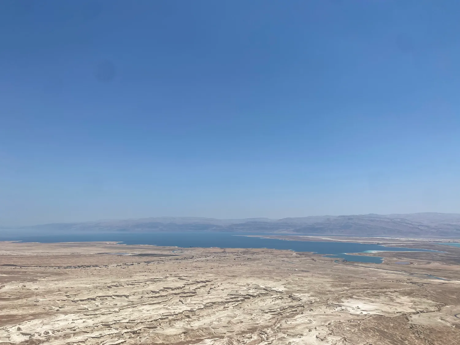 A wide view over a pale, cracked desert plain stretching to blue water, with bare hills along the far shore and a clear sky above.
