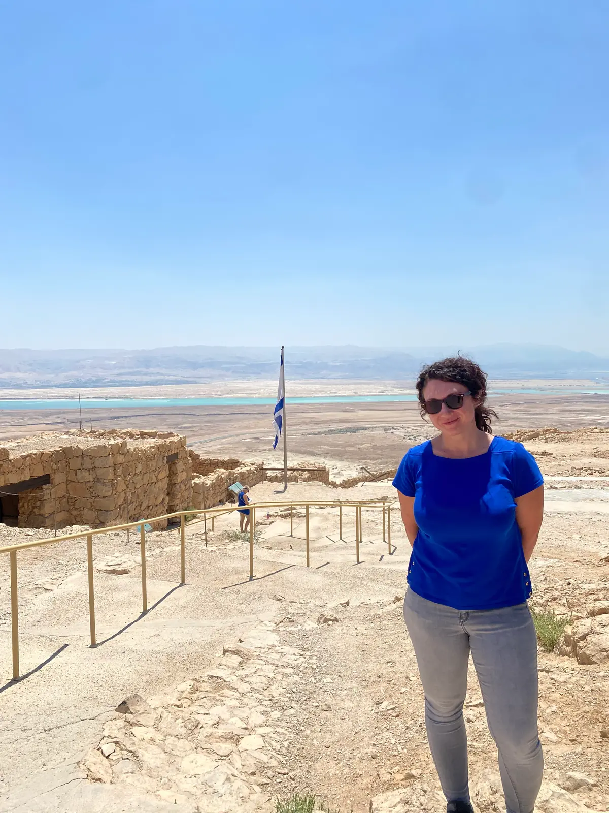A woman in a blue shirt and dark glasses standing on a stone path at a fortress site, a flag on a pole beside her and the pale plain and blue water far below her.