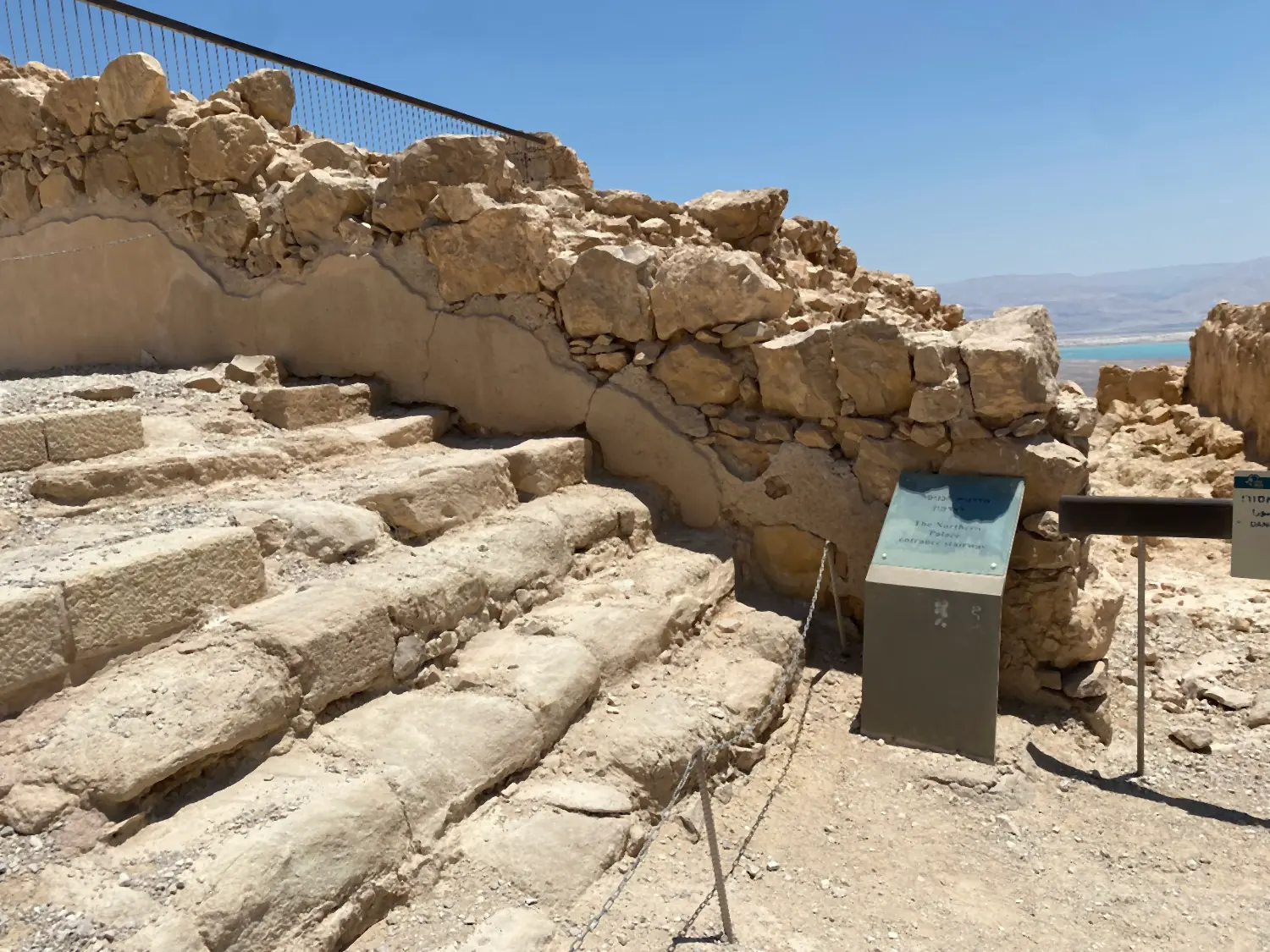 Worn stone steps running up beside a wall of rough rock, a black railing along the top of the wall and an interpretive panel standing at its foot.