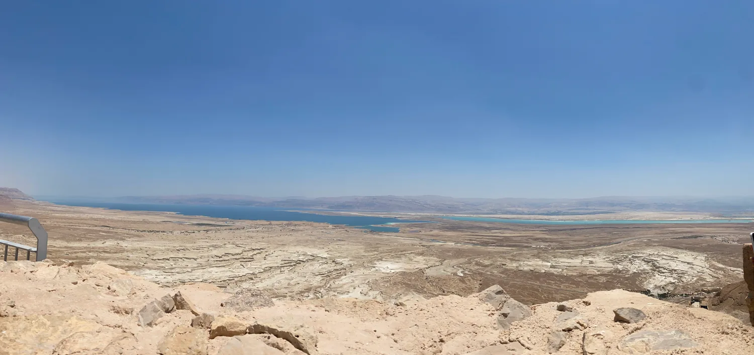 A long, low view over pale desert ground to a strip of blue water, bare hills along the horizon and rough rock in the foreground.
