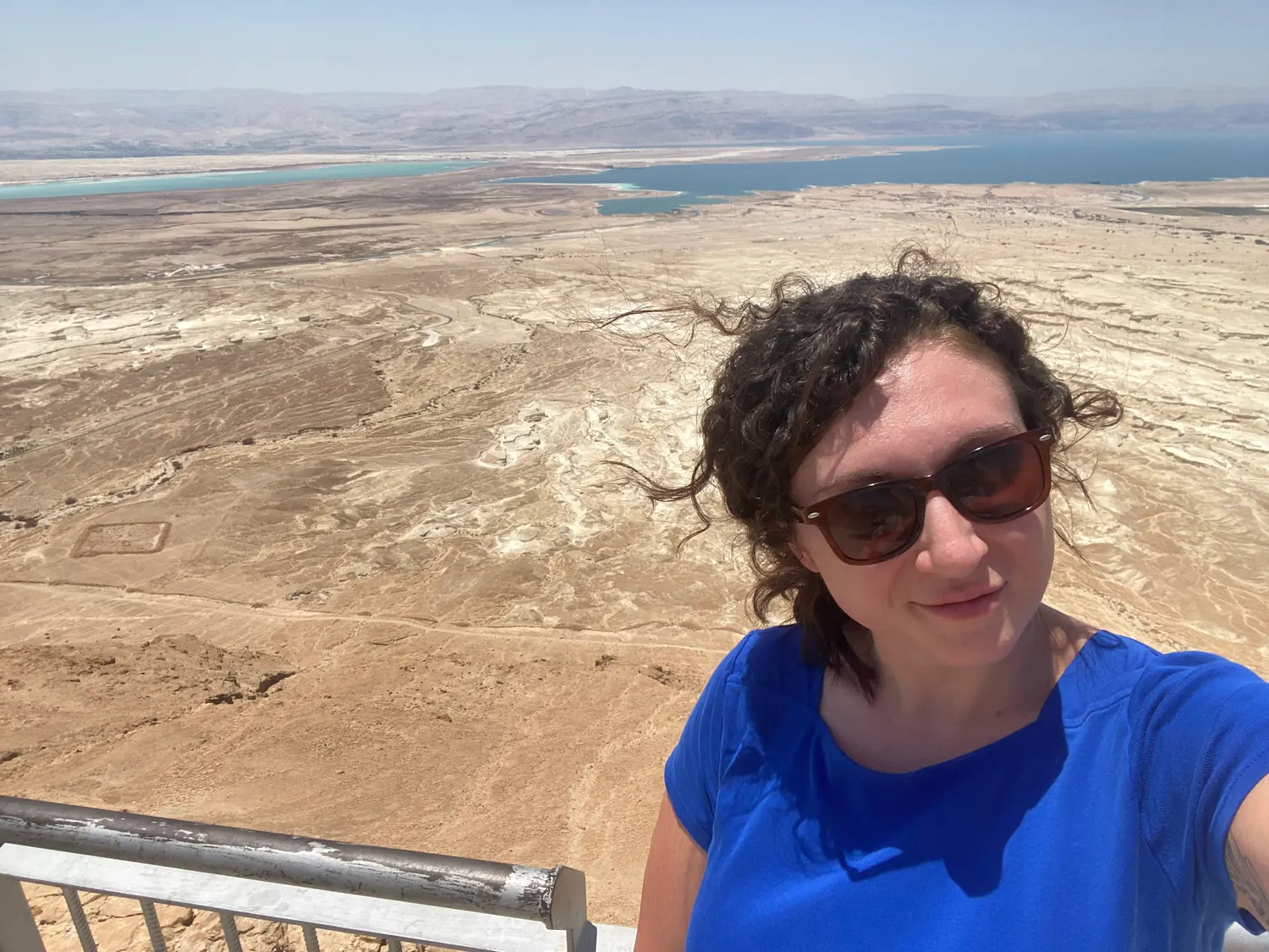 A selfie of a woman in a blue shirt with the pale desert plain and blue water spread out behind her, a metal railing below.