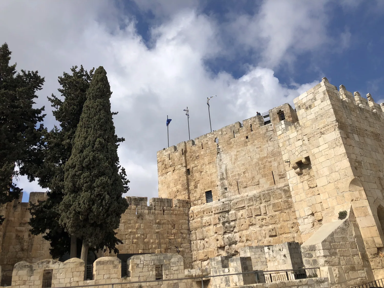 A long front of pale stone blocks with cypress trees growing above it, a flag on a pole at the top and rough rocks along the foot of the wall.