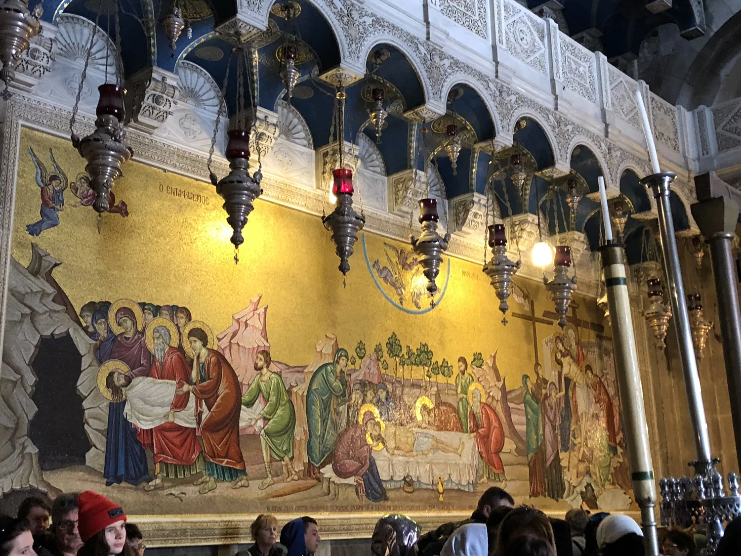 A long gilded wall covered in painted figures behind a row of hanging lamps, with the tops of visitors' heads along the bottom edge.