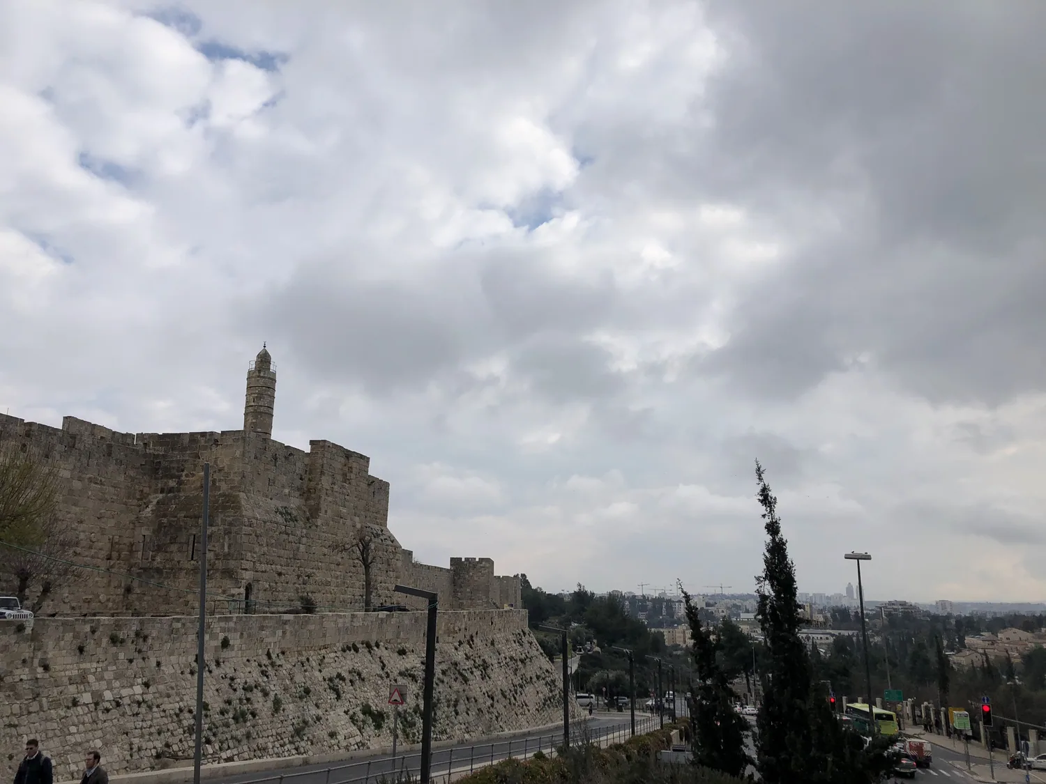 A long stone city wall with a tall tower behind it running along the far side of a road, cypress trees in front of it and a wide cloudy sky above.