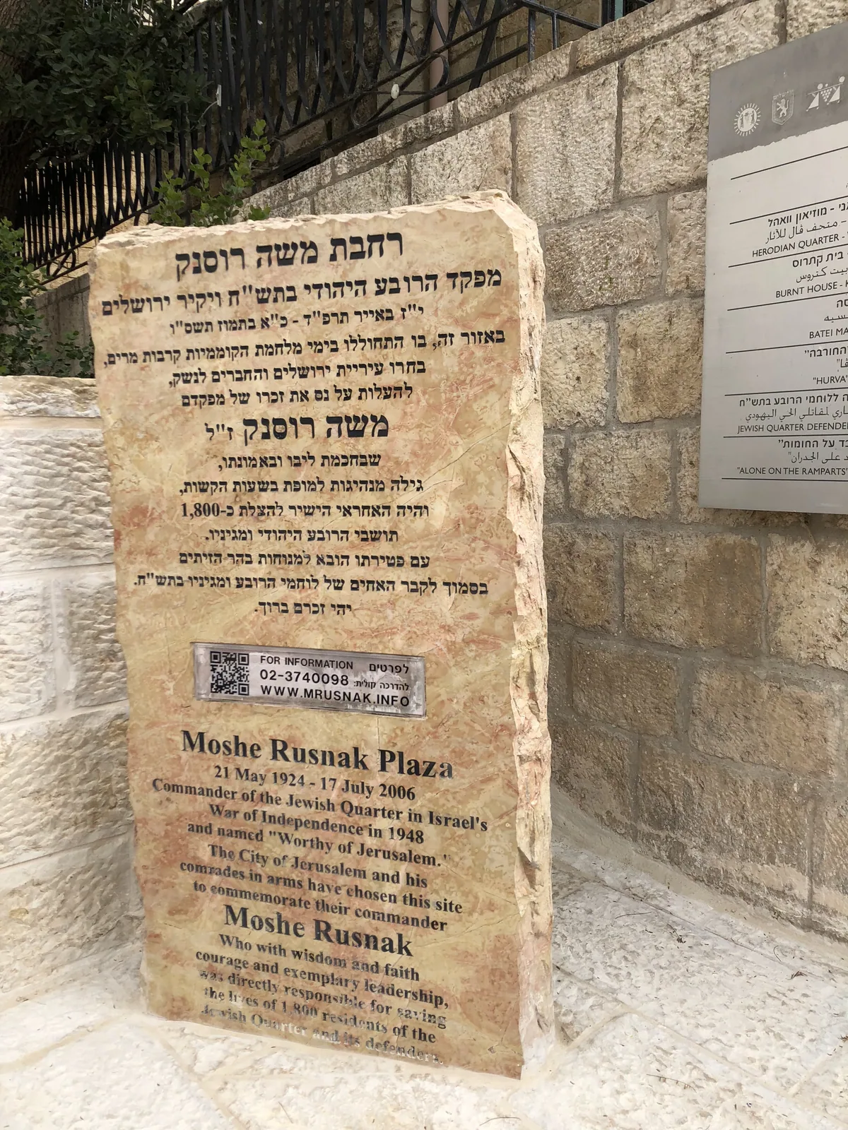 A tall carved stone panel standing against a stone wall, its face covered in dense lettering, with a smaller panel fixed to the wall beside it.