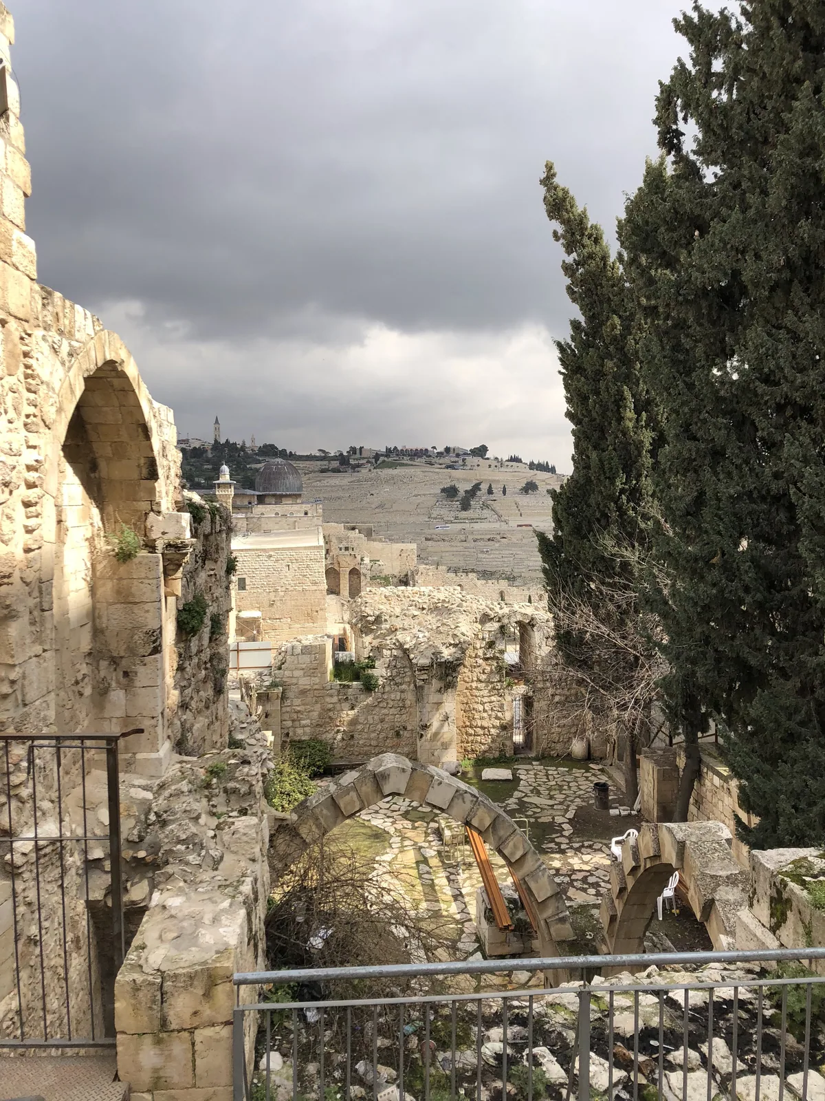 A view over old stone buildings and wall tops from a raised path, cypress trees along the ridge and a metal railing across the front.