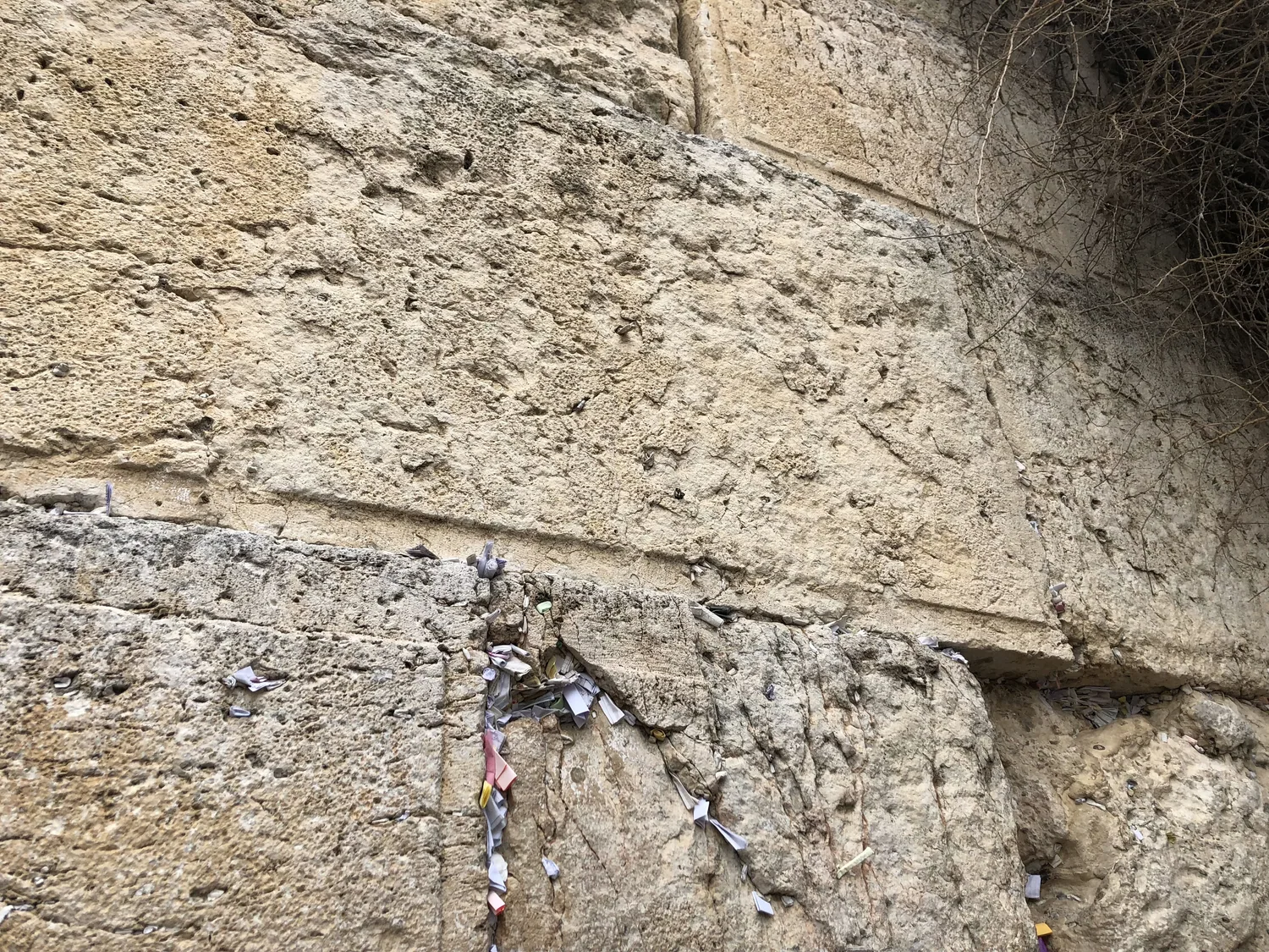 A close view of a huge wall of weathered stone blocks with small folded paper notes pushed into the gaps between them.