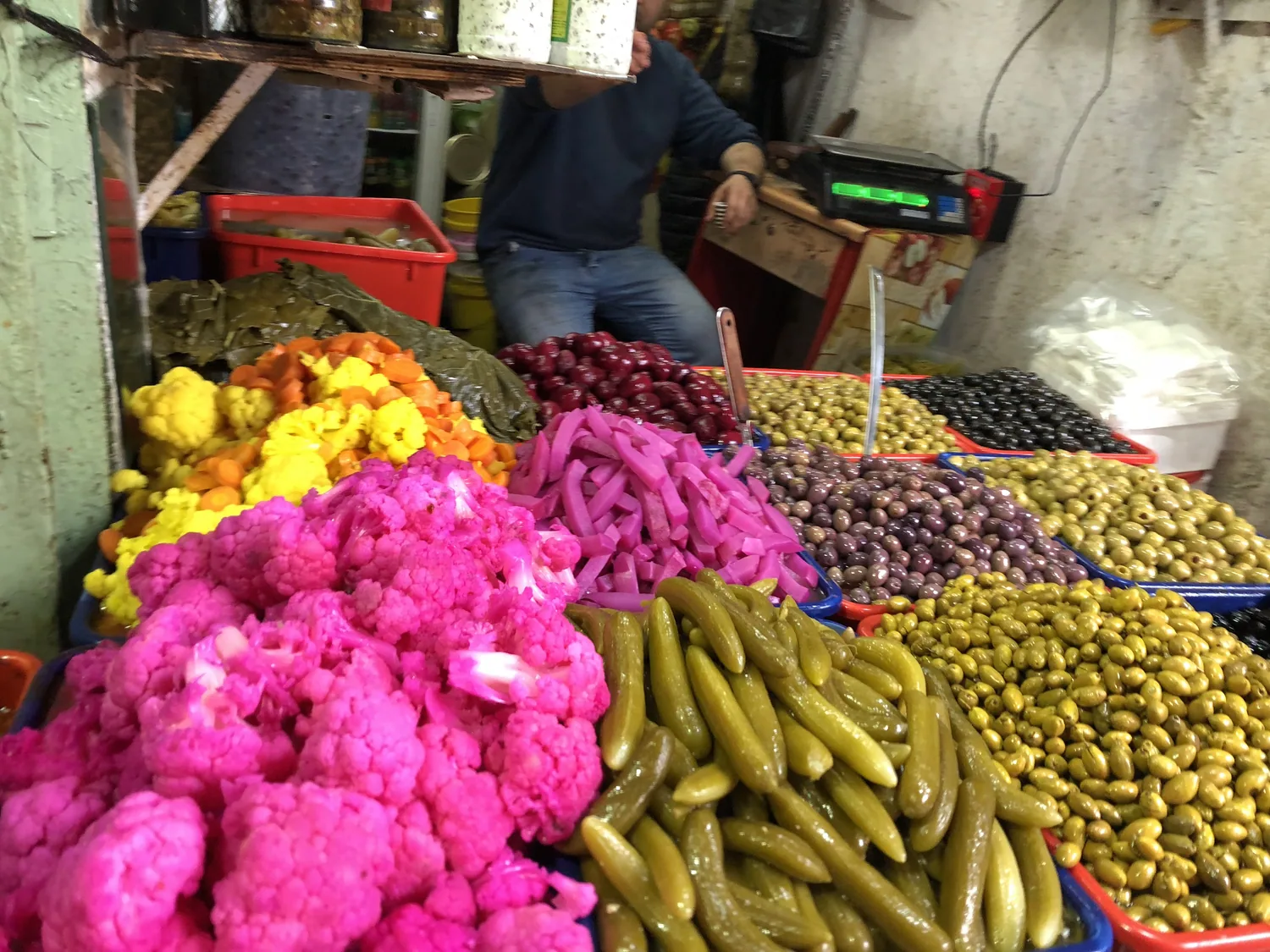 A stall counter covered in trays of pickled vegetables and olives in bright pinks, greens and yellows, with a scale at one end.