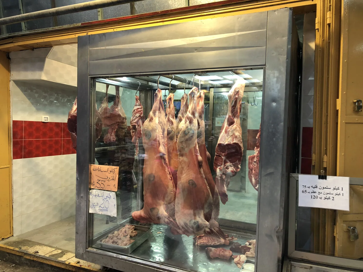 A shop window with cuts of meat hanging on hooks behind the glass, small cards taped to the pane and tiles on the wall behind.
