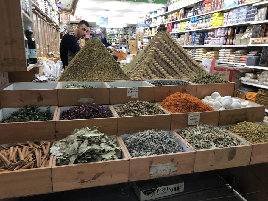 Wooden boxes of ground spices and dried herbs heaped high in a shop doorway, packets stacked on shelves behind and a man standing at the counter.