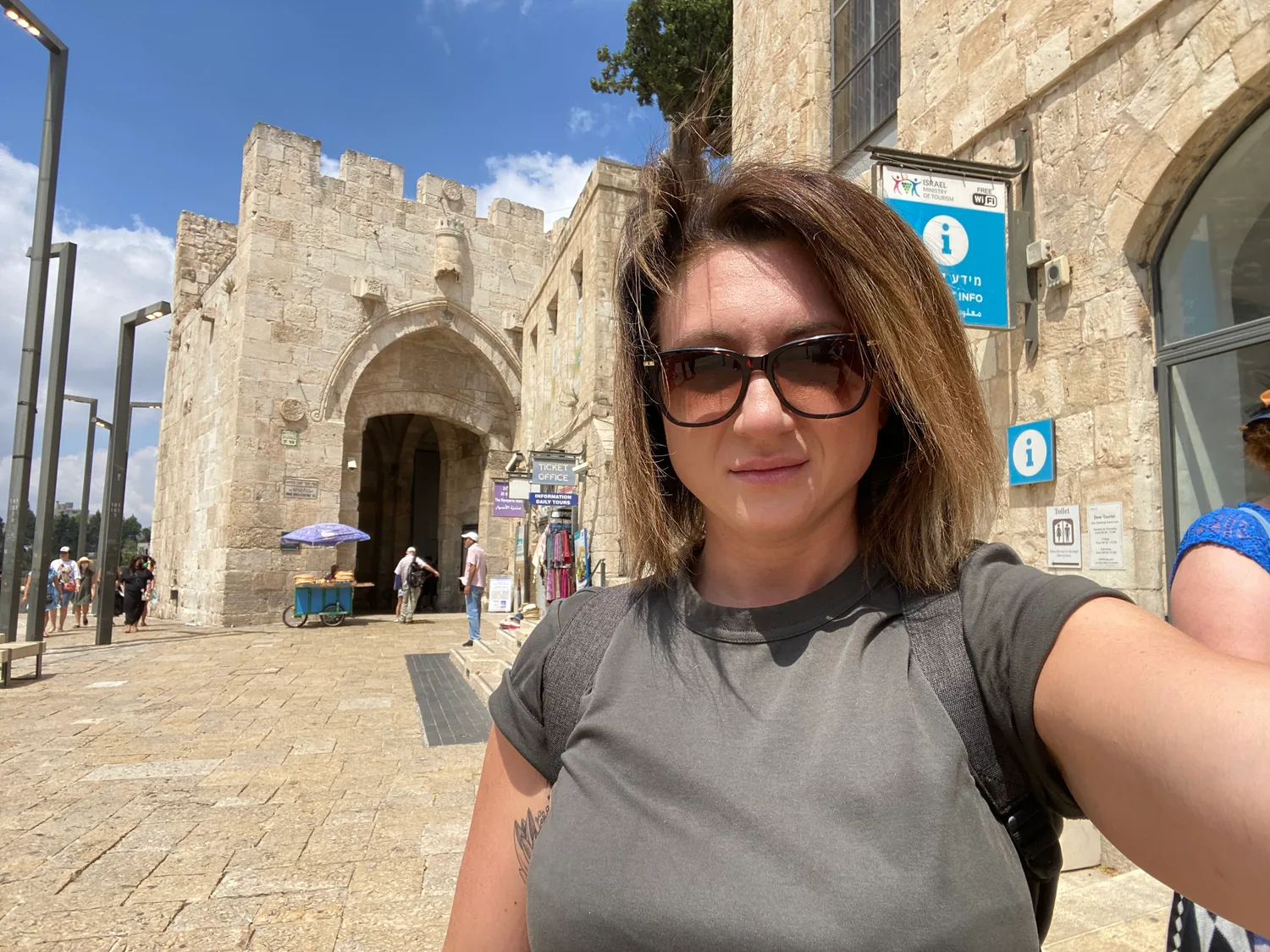 A selfie of a woman in a grey top and dark glasses in front of the stone gate of an old walled city in bright sun, the gate's crenellated top and pointed arch behind her, a blue information sign on the wall to her right with a smaller sign and a toilet sign below it, a ticket office sign at the corner of the building, and other visitors on the paved plaza.