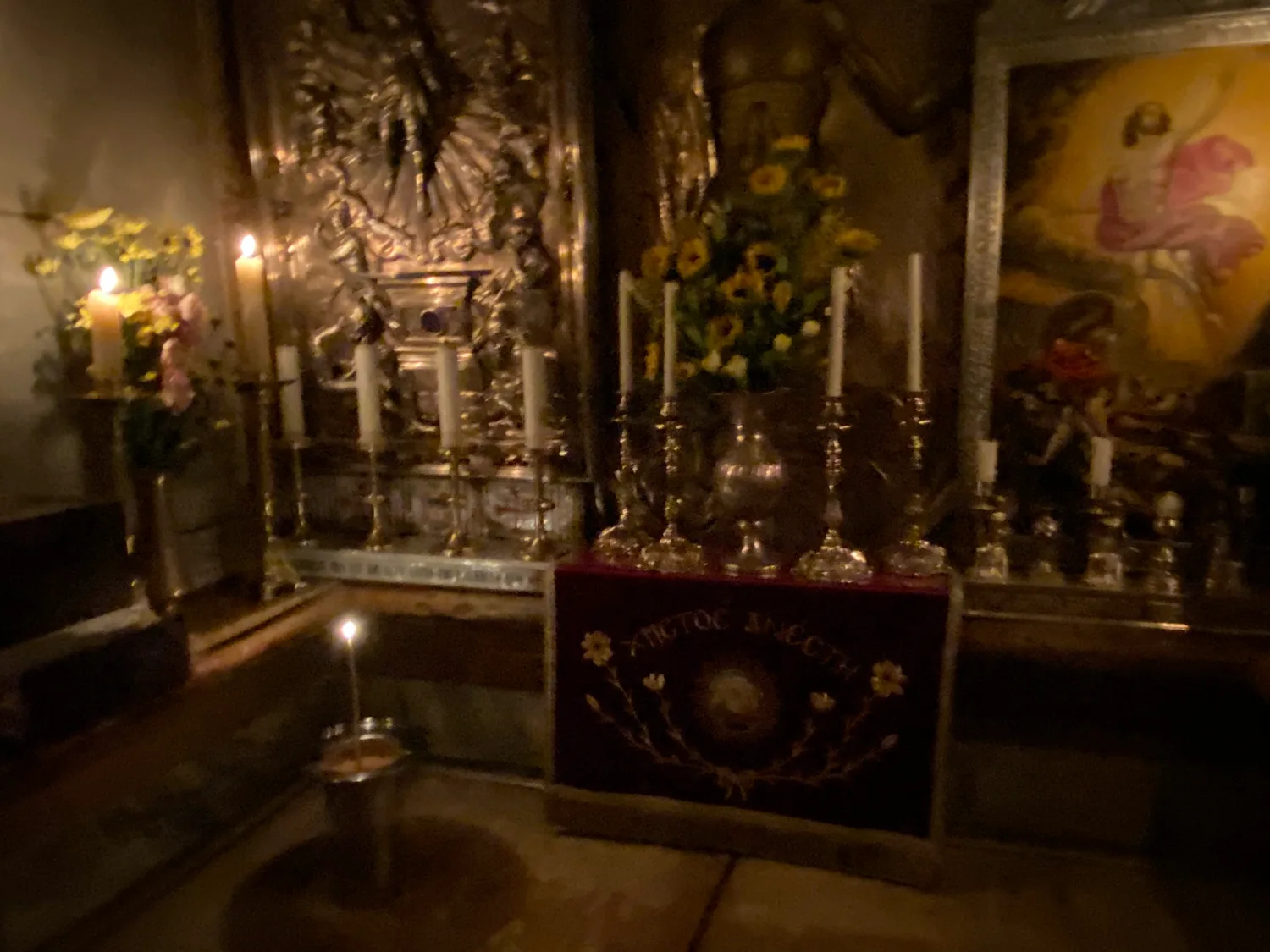 A dim chapel interior with gold-framed painted panels along the wall, silver candle stands, lamps lit in front of a red and gold hanging and stone underfoot.