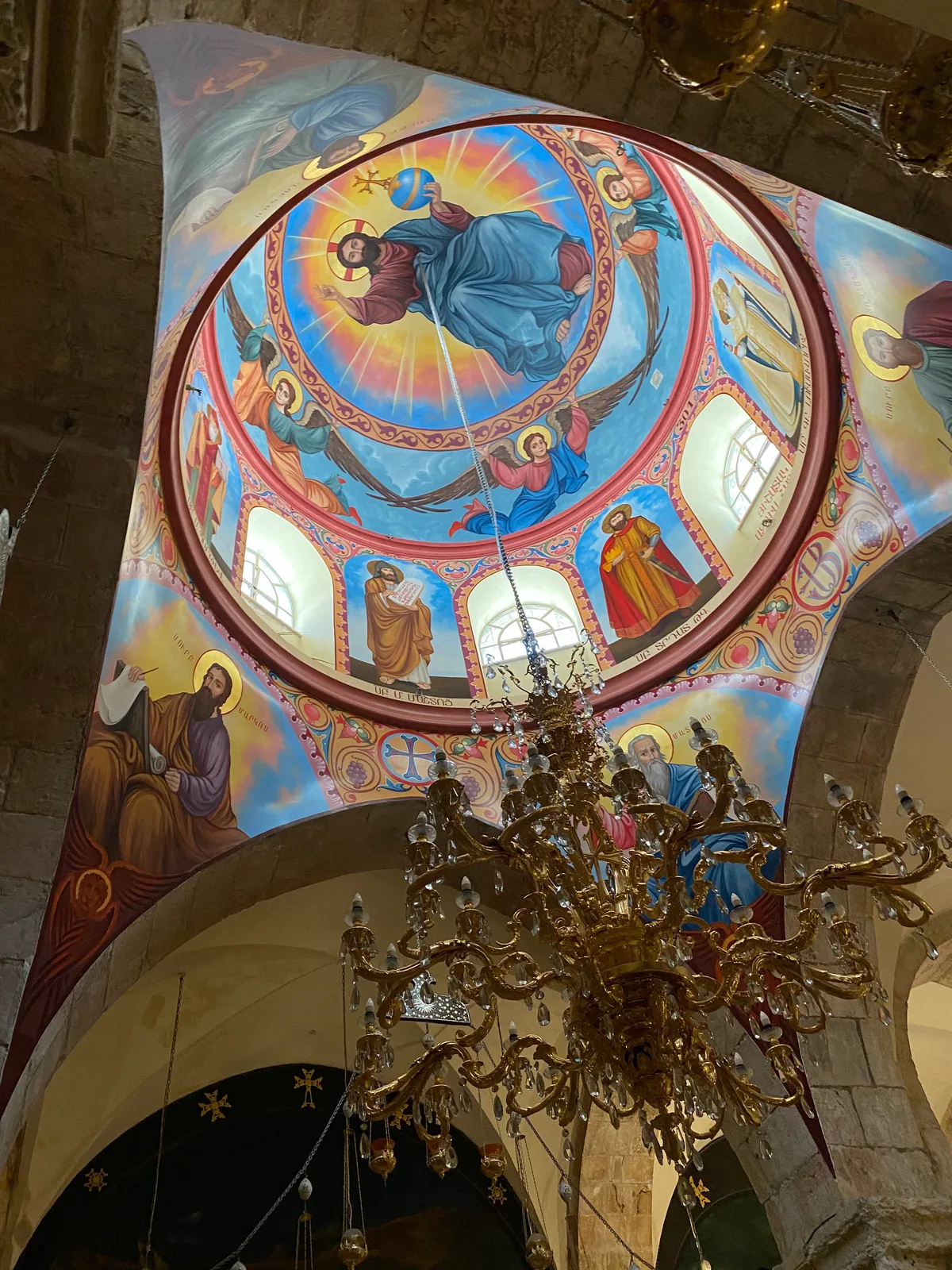 An arched ceiling painted with figures around a circle of windows, seen from below with a large brass chandelier hanging on a chain in front of it.