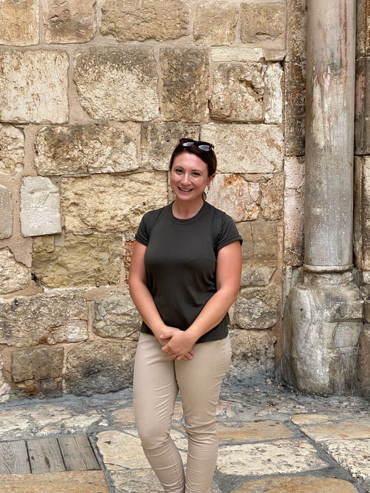 A woman in a grey top and light trousers standing against a wall of old stone blocks, smiling, with a worn stone column at the side and stone paving underfoot.