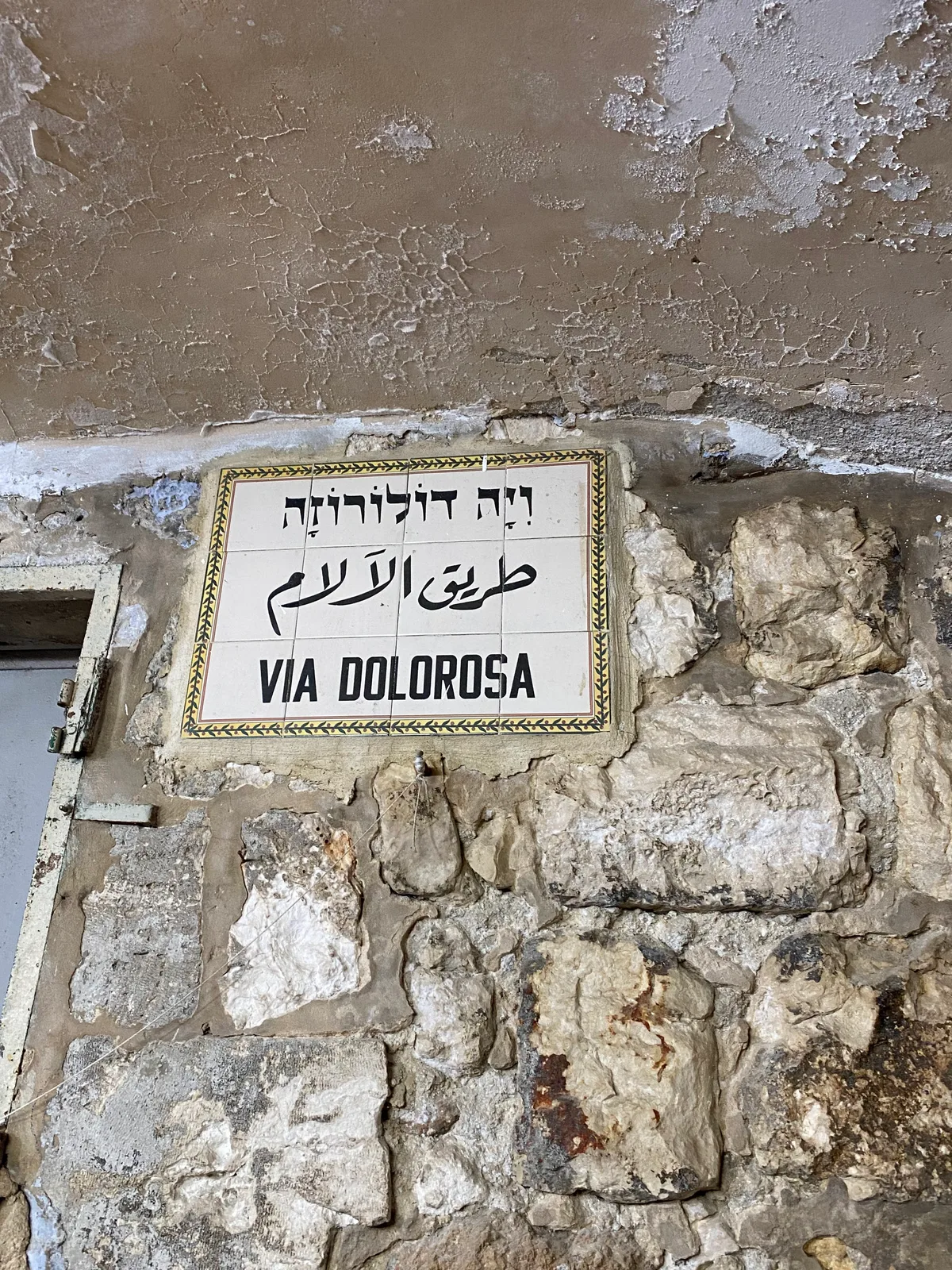 A tiled street sign set into an old stone wall with its lettering in three scripts, a patterned tile border around the writing and a metal door frame at the left.
