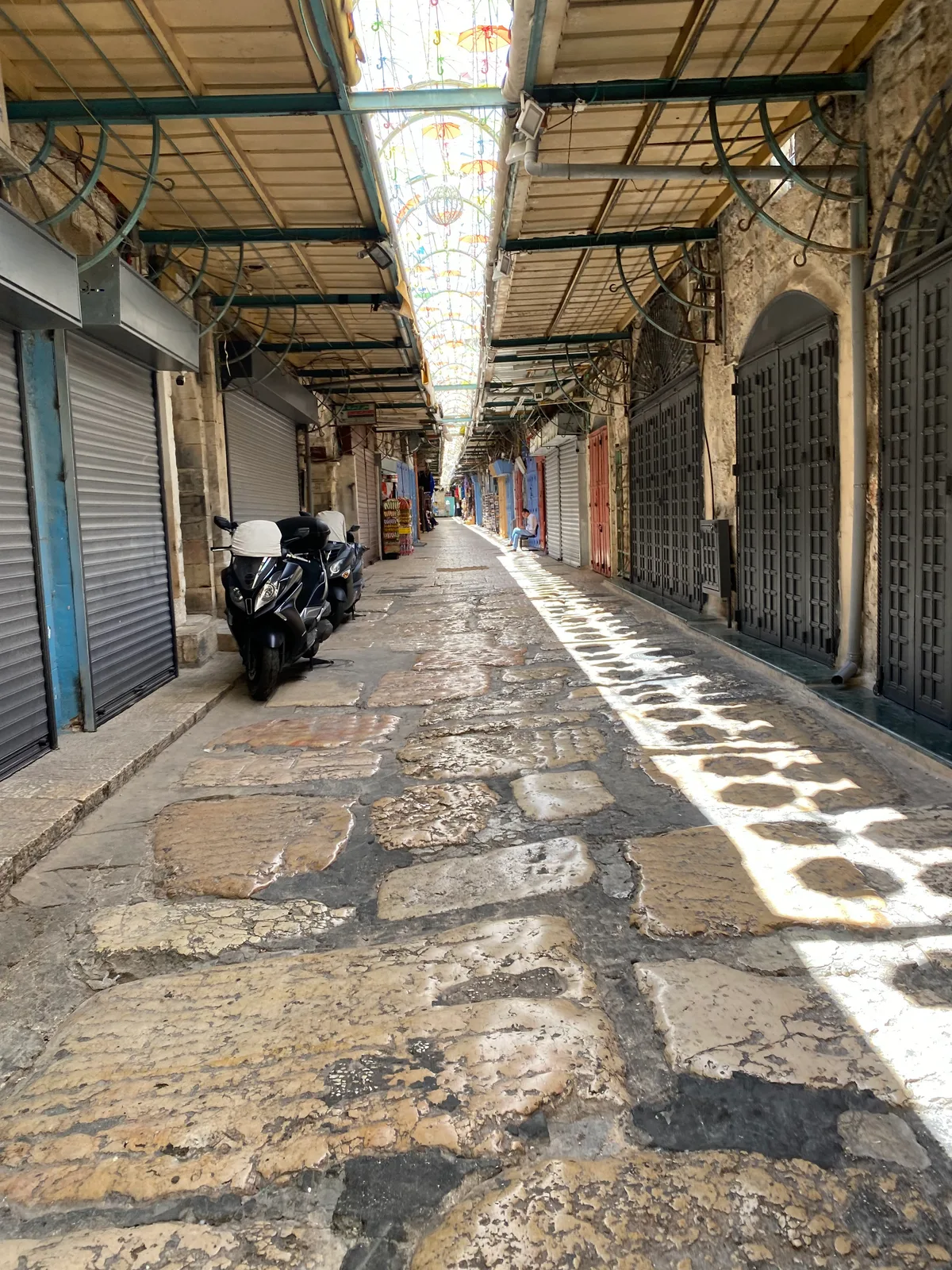 A covered stone lane running away between buildings with their shutters down, coloured umbrellas hanging overhead, motorcycles parked along the paving and light falling through in stripes.