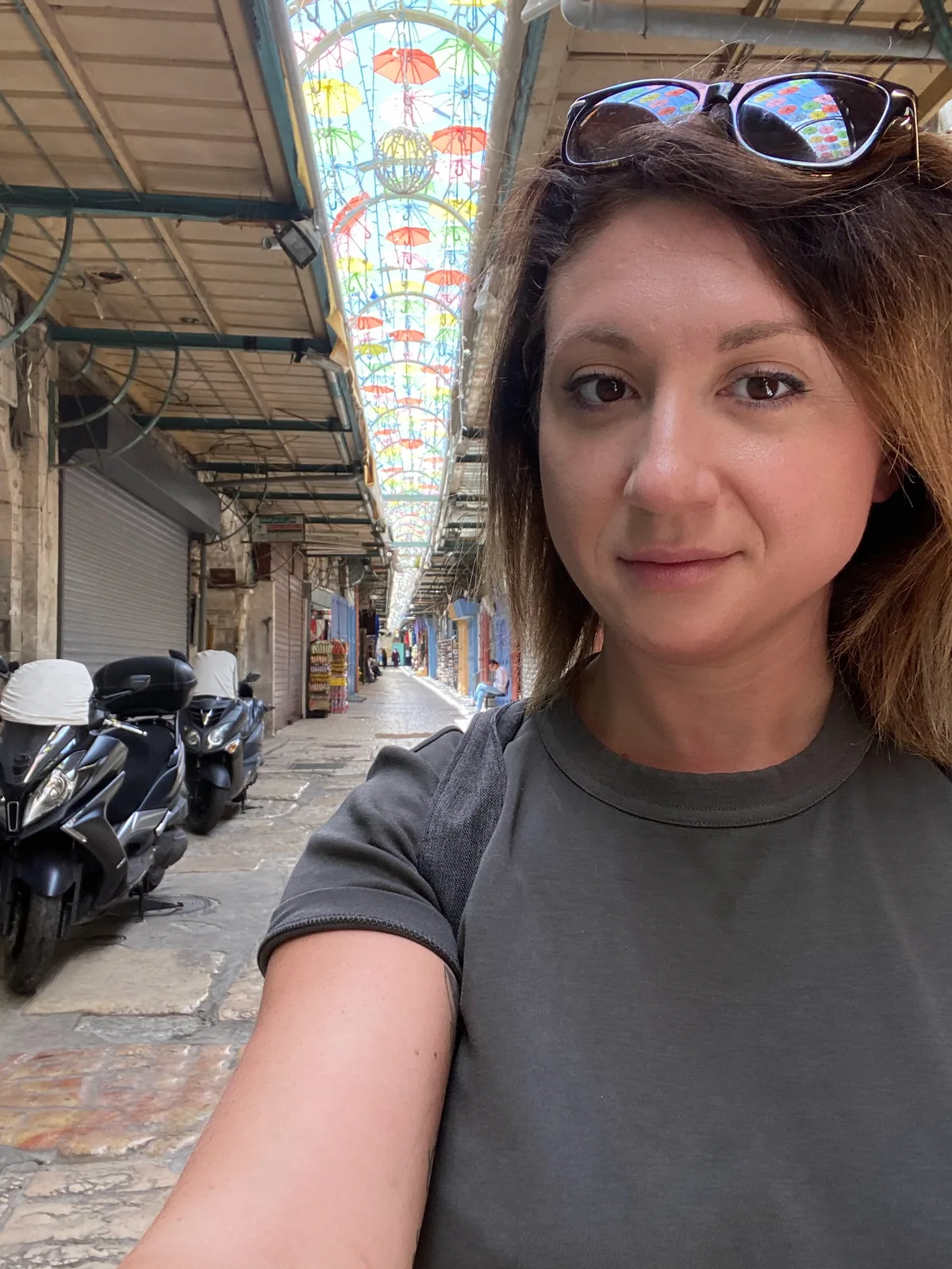 A selfie of a woman in a grey top with glasses pushed up on her head, standing in a stone lane with coloured umbrellas hanging overhead and motorcycles parked along the side.