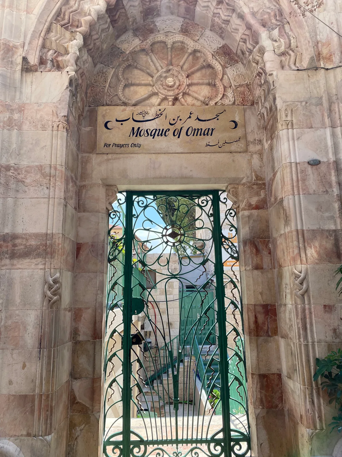 A stone doorway with lettering carved along the top and a sign panel above it, an open iron gate below and a wall of patterned stone around it.