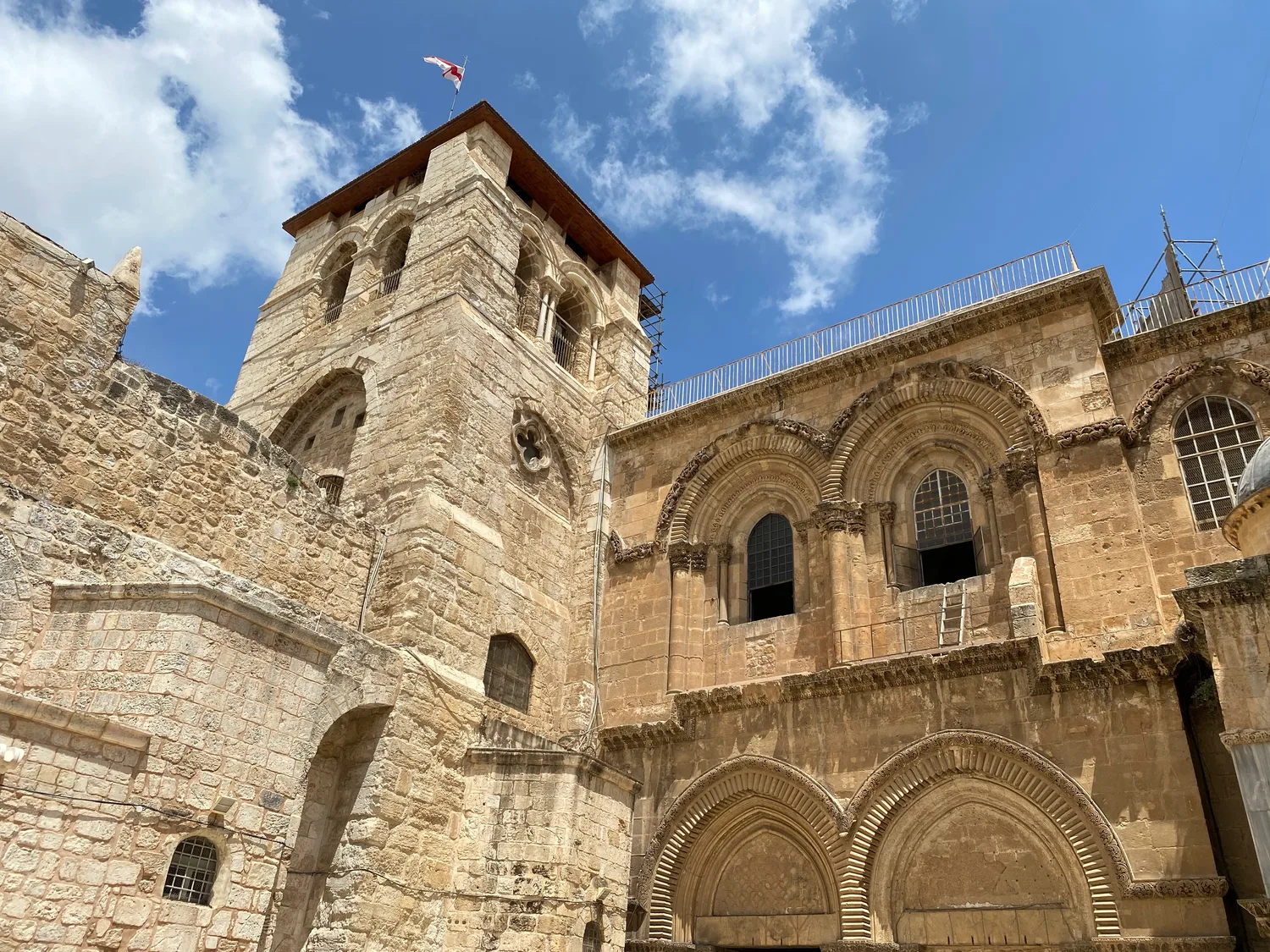 A square stone tower with a flag flying above it standing beside a pale stone front with paired arched windows, under a blue sky with white cloud.