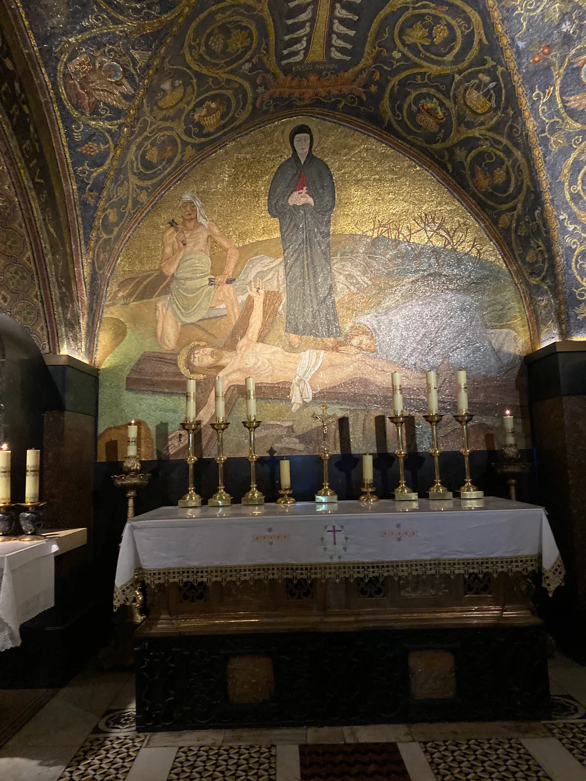 A small stone chapel with a gold mosaic above the altar, tall candlesticks along it, a white cloth over the front and patterned tiles on the floor.