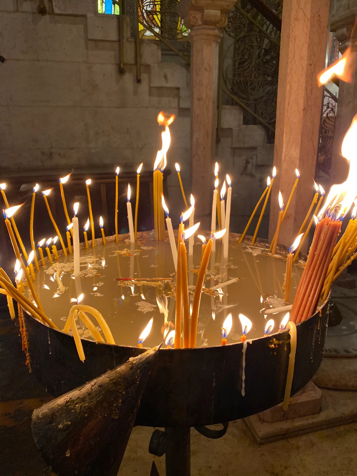A round metal stand full of lit candles set in sand, the flames crowded together and wax running down the metal side.
