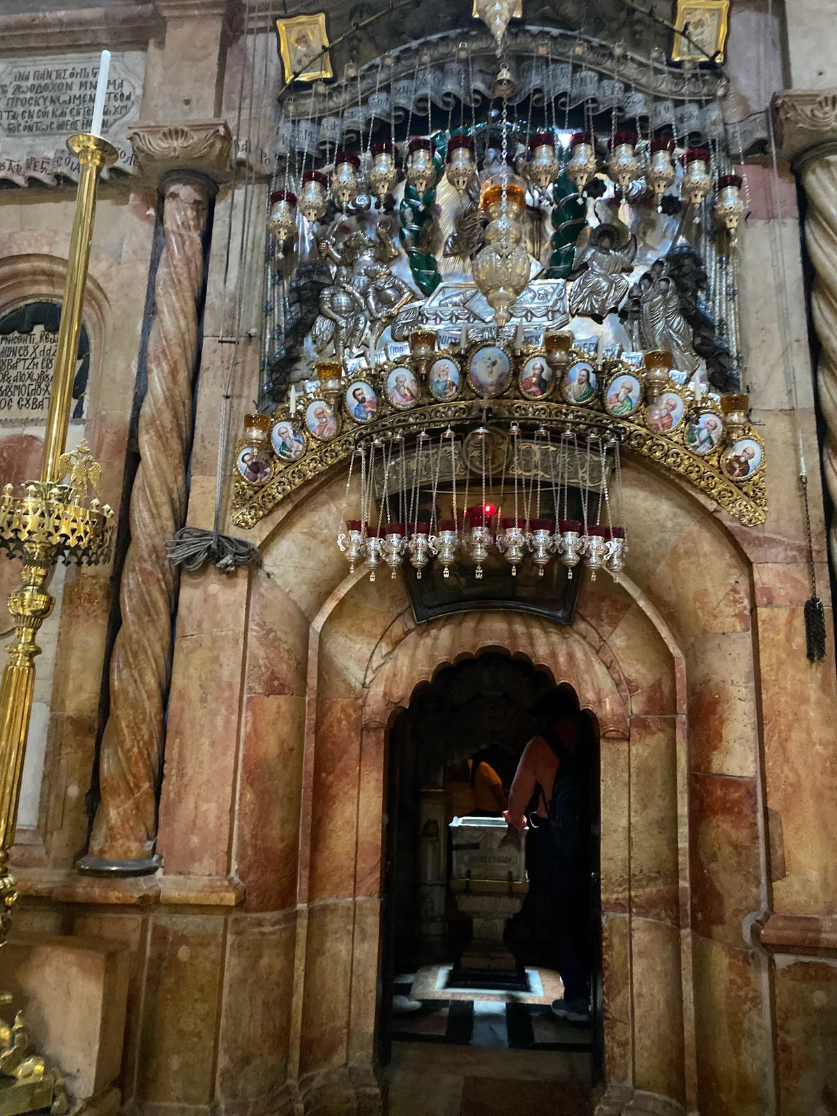 An ornate stone entrance with a cluster of hanging metal lamps and small painted plaques over the arch, columns on either side and a dark doorway below.