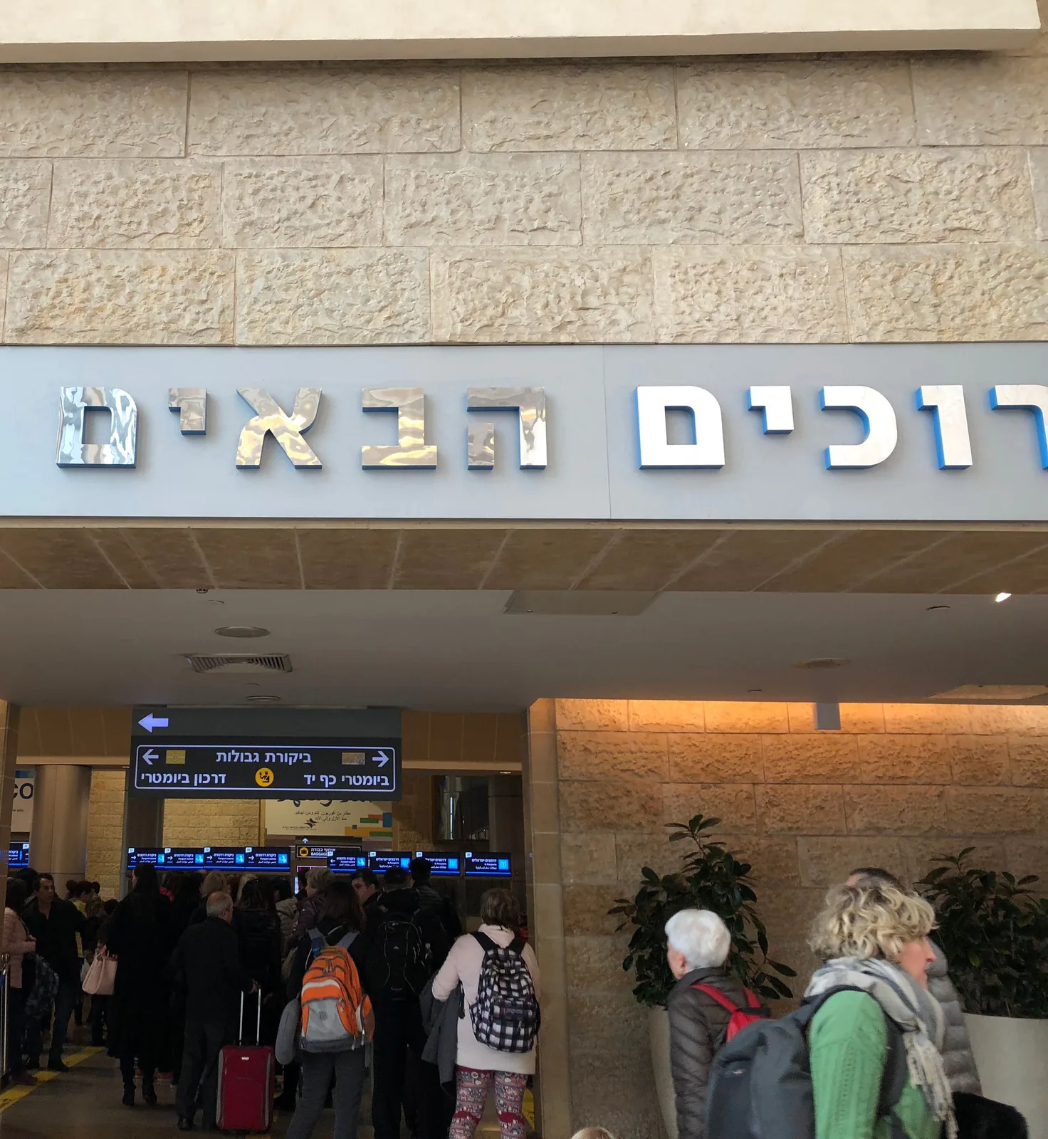 An arrivals hall with a wide lettered sign above a row of lit counters, travellers with luggage walking through and small groups waiting.