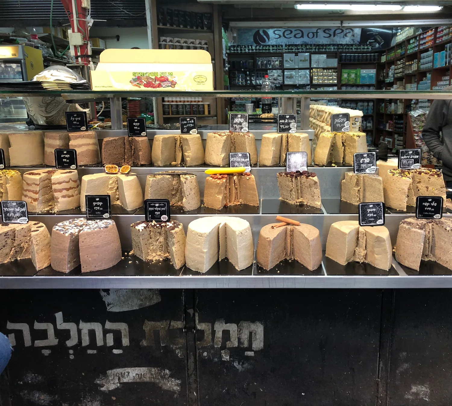 A cheese counter stacked with wheels and cut pieces of cheese, a lit display case behind it and a lettered shop board on the wall above.
