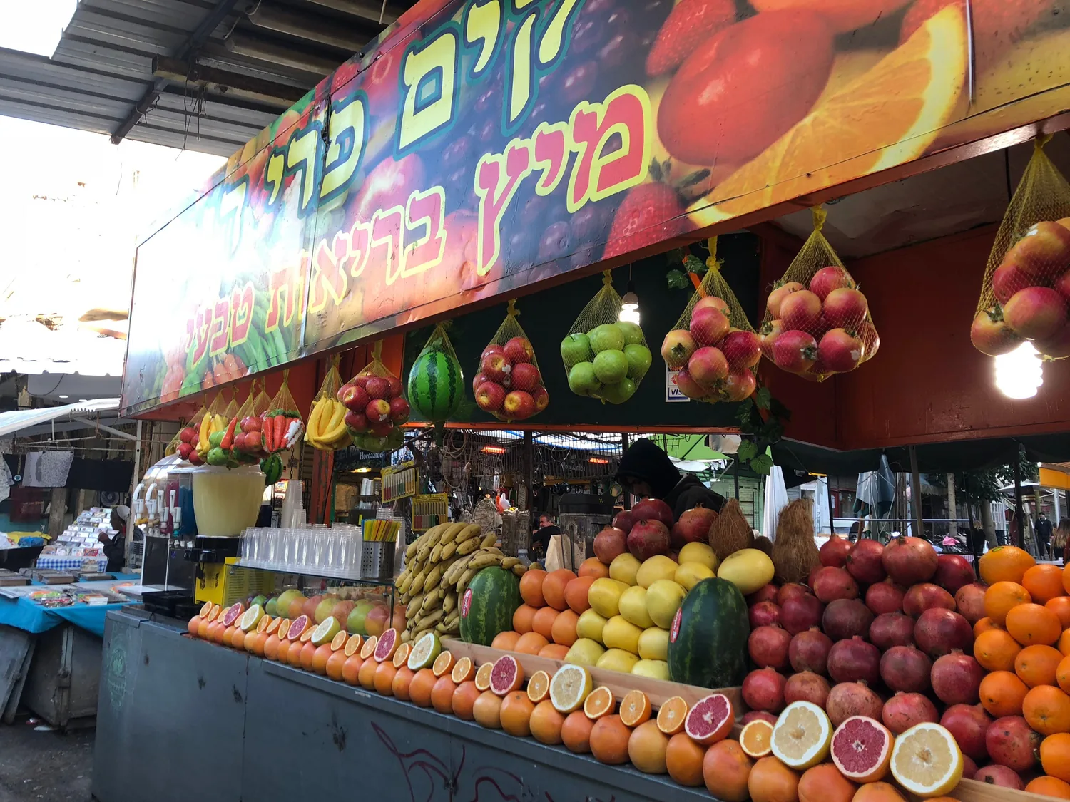 A market fruit stall with oranges, melons and bananas stacked in tiers below a painted board, and bags of produce hanging above them.