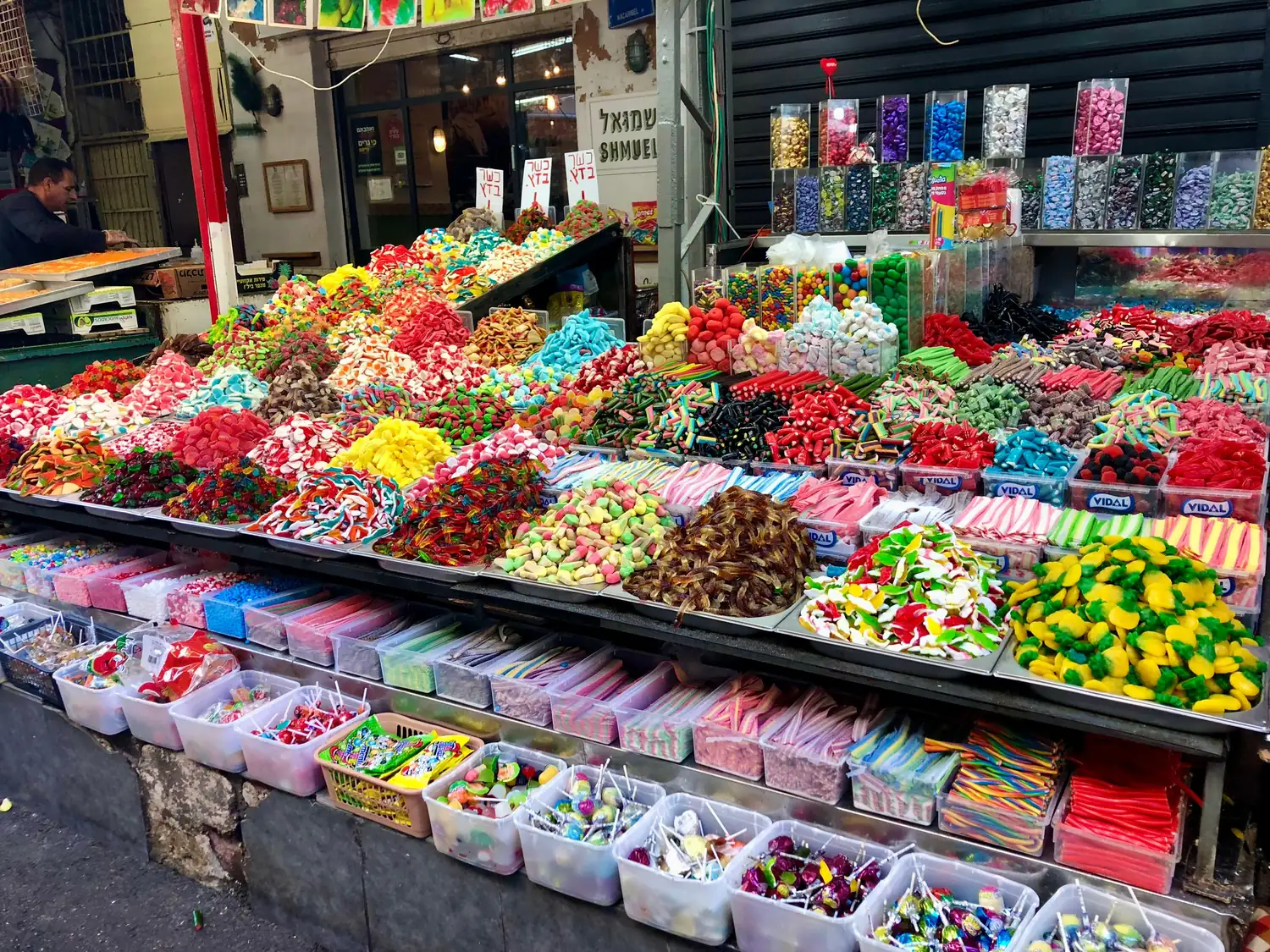 A market stall of sweets, trays of brightly coloured candies and jelly pieces in rows and boxes of more sweets stacked below them.