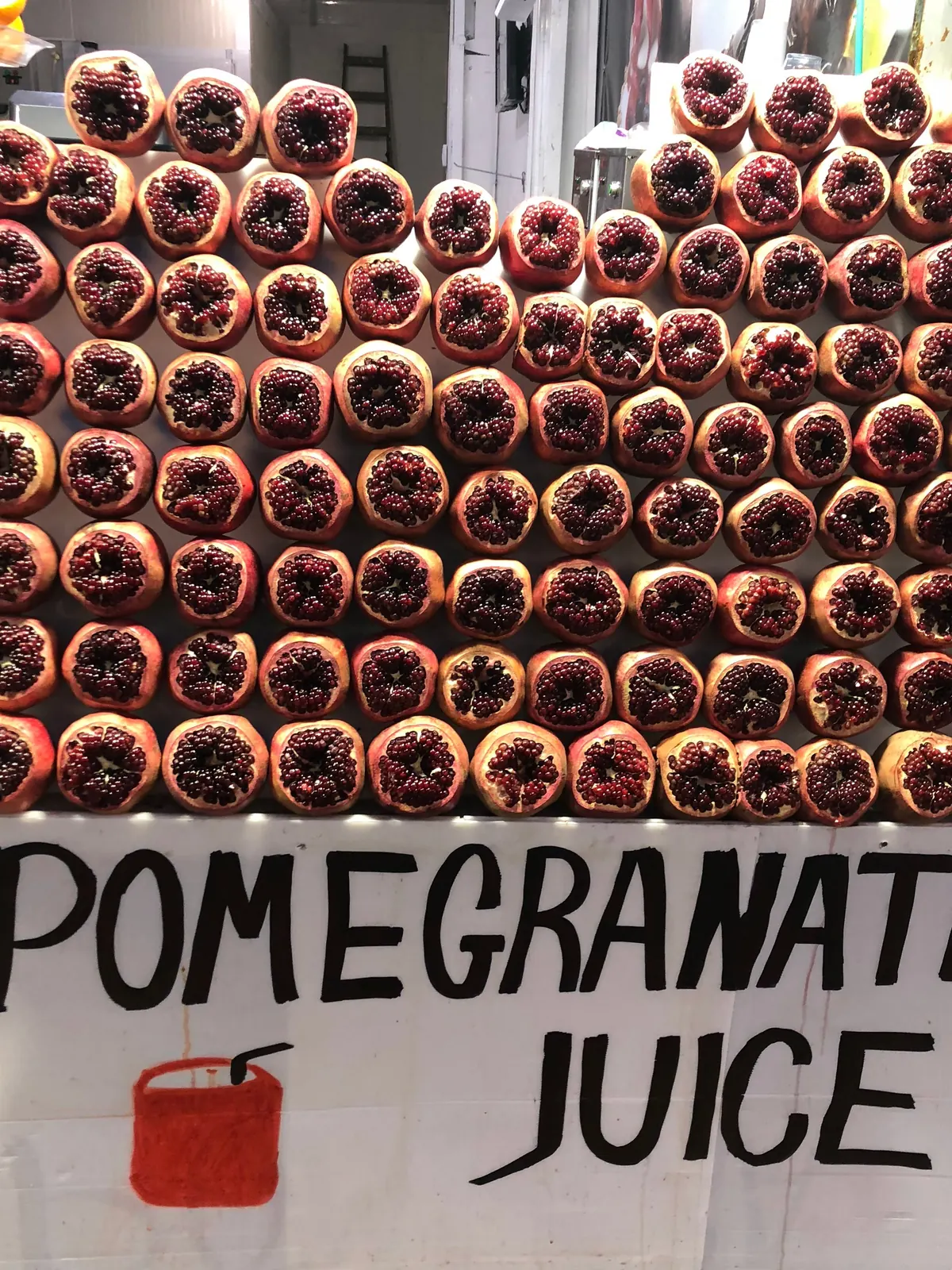 A stall stacked high with pomegranates against a painted board, with a smaller painted sign below the fruit.