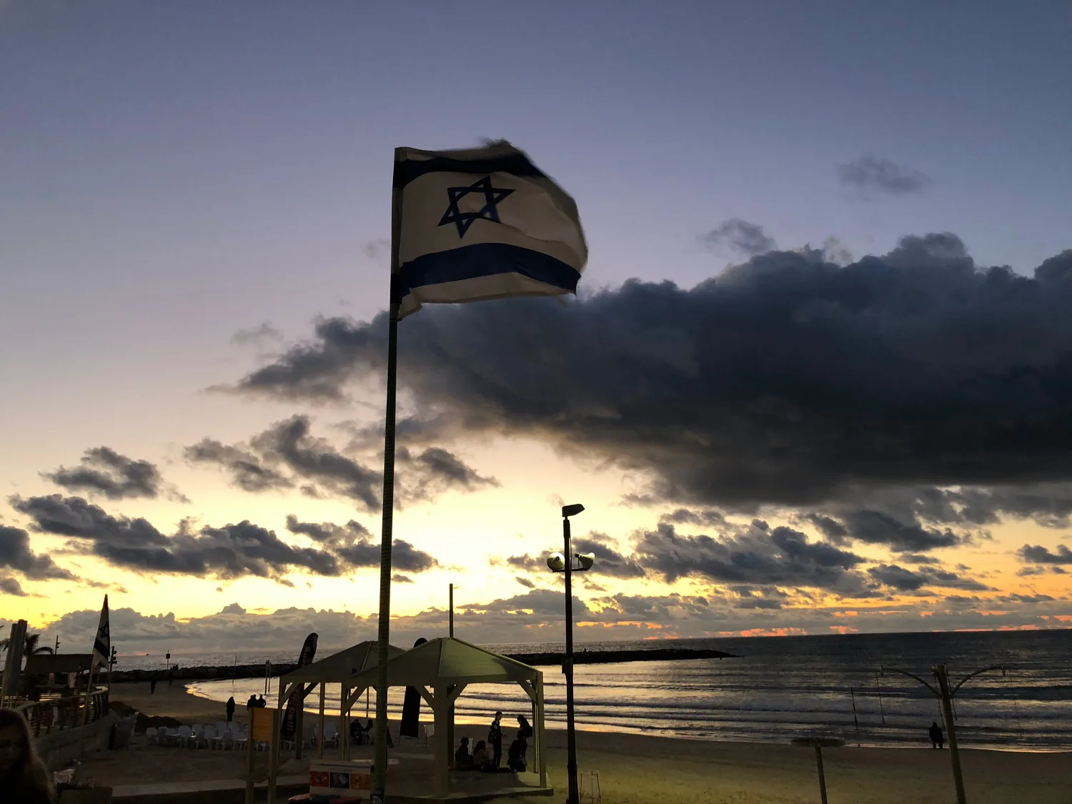 A flag on a tall pole above a beach at sunset, a wide band of cloud across the sky and people sitting on the sand below.