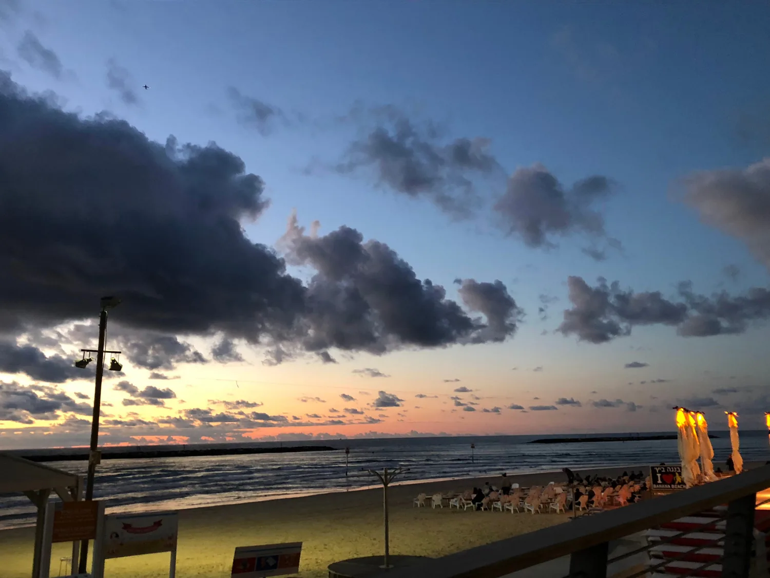 A beach at dusk seen from a promenade, the sand and the sea below, a bar's lit umbrellas and tables along the front and a band of cloud over the water.