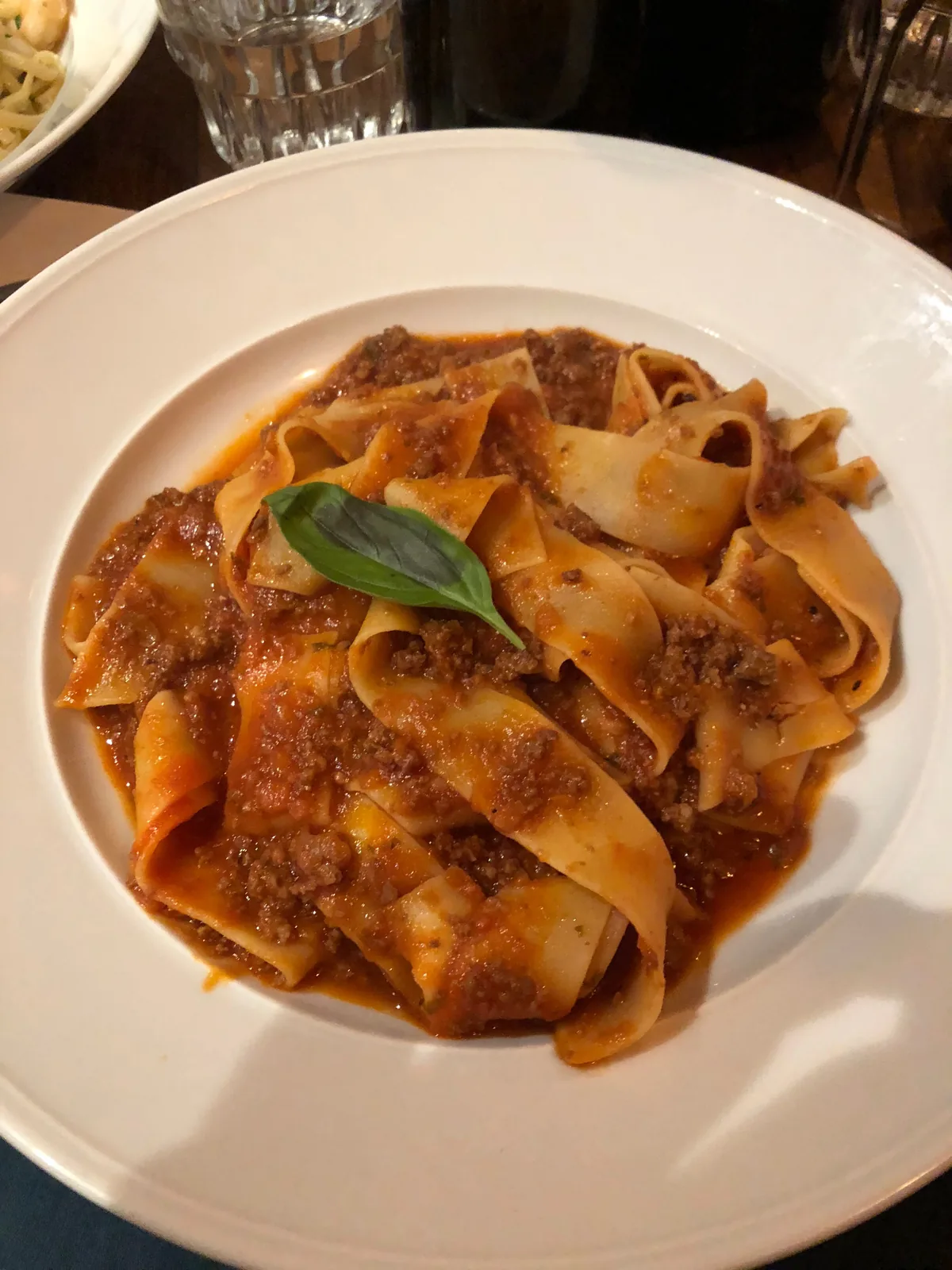 A white plate of ribbon pasta in sauce on a table, with a leaf of basil on top.