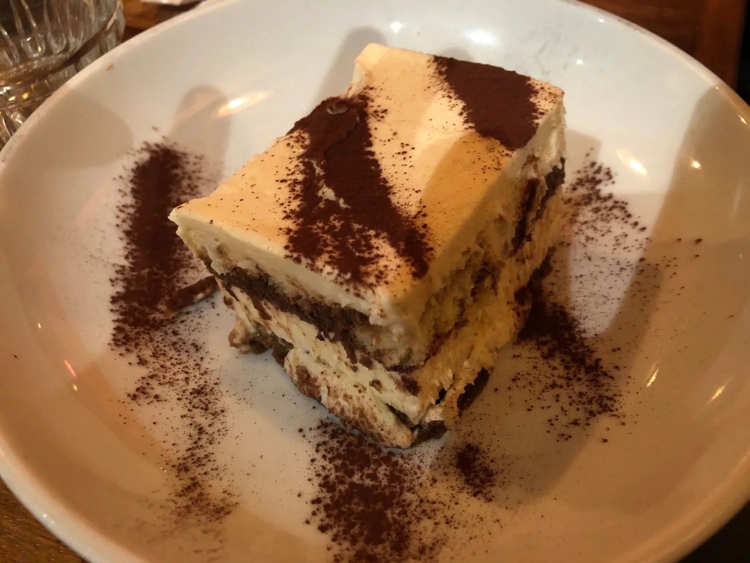 A slice of layered cake on a white plate, cocoa dusted over the plate around it and cream on the top.