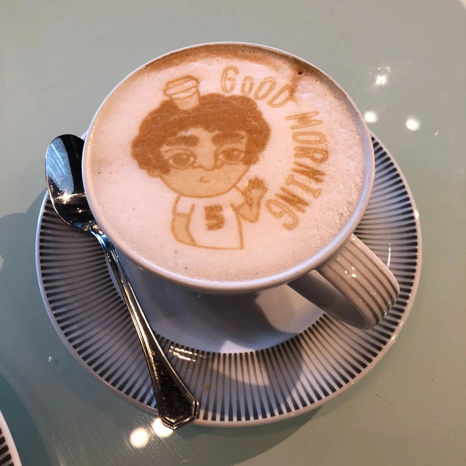A cup of coffee on a saucer with a cartoon face drawn in the foam and lettering beside it, a spoon resting on the saucer.