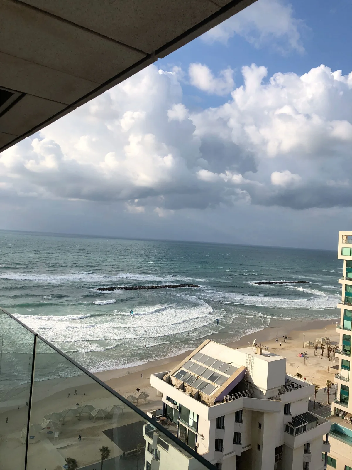 The sea and a beach seen from a balcony, the balcony's glass down one side, waves breaking in rows toward the sand and low white buildings along the shore.